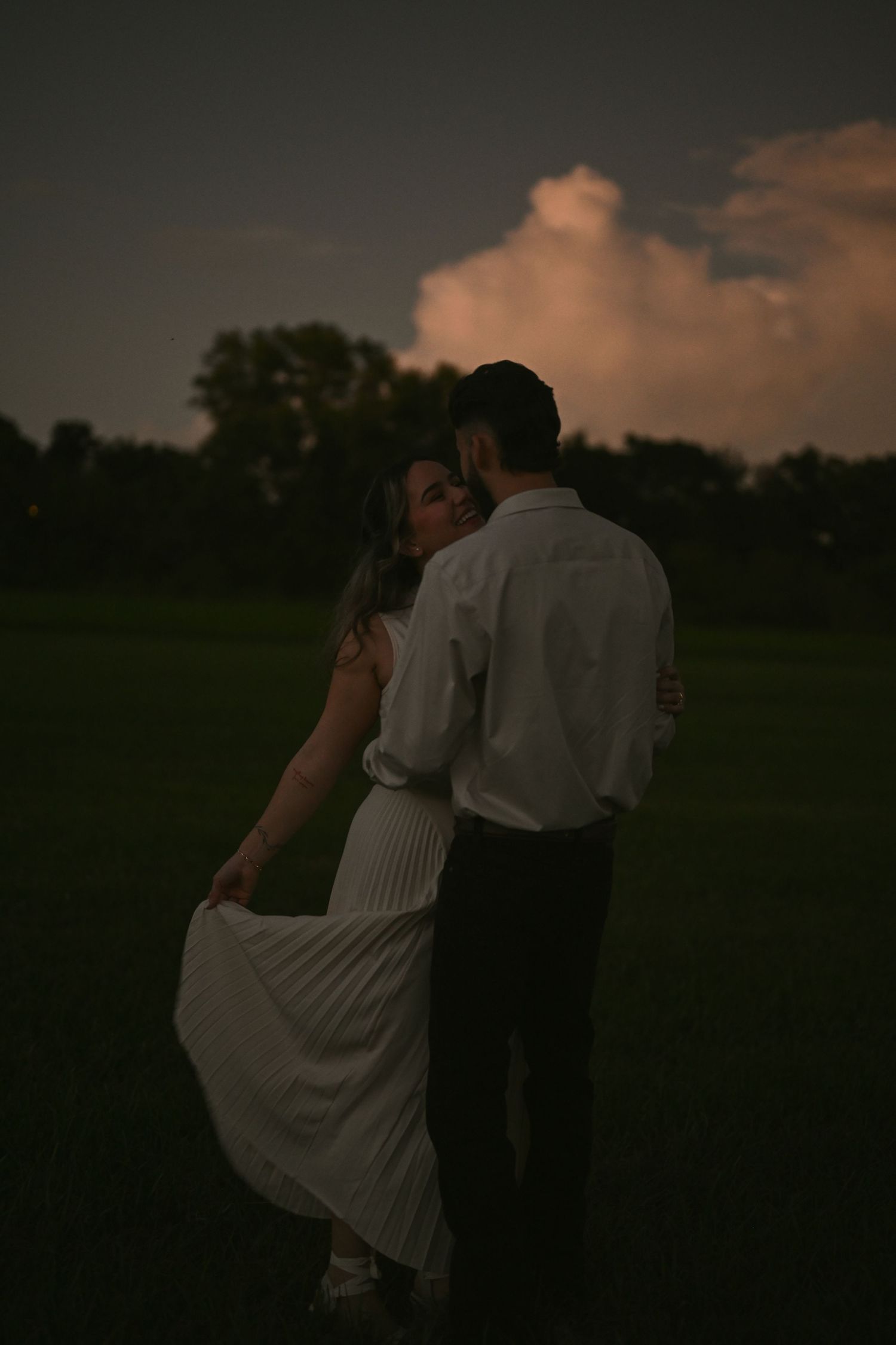 Couple embracing romantically at dusk in a green field, woman in white pleated dress, dramatic cloudy sky backdrop.