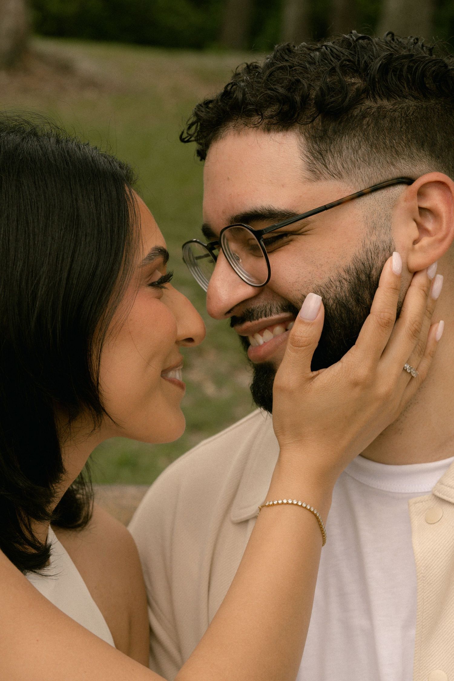 A close romantic moment as a smiling woman gently cups a bearded mans face while nearly touching noses outdoors.