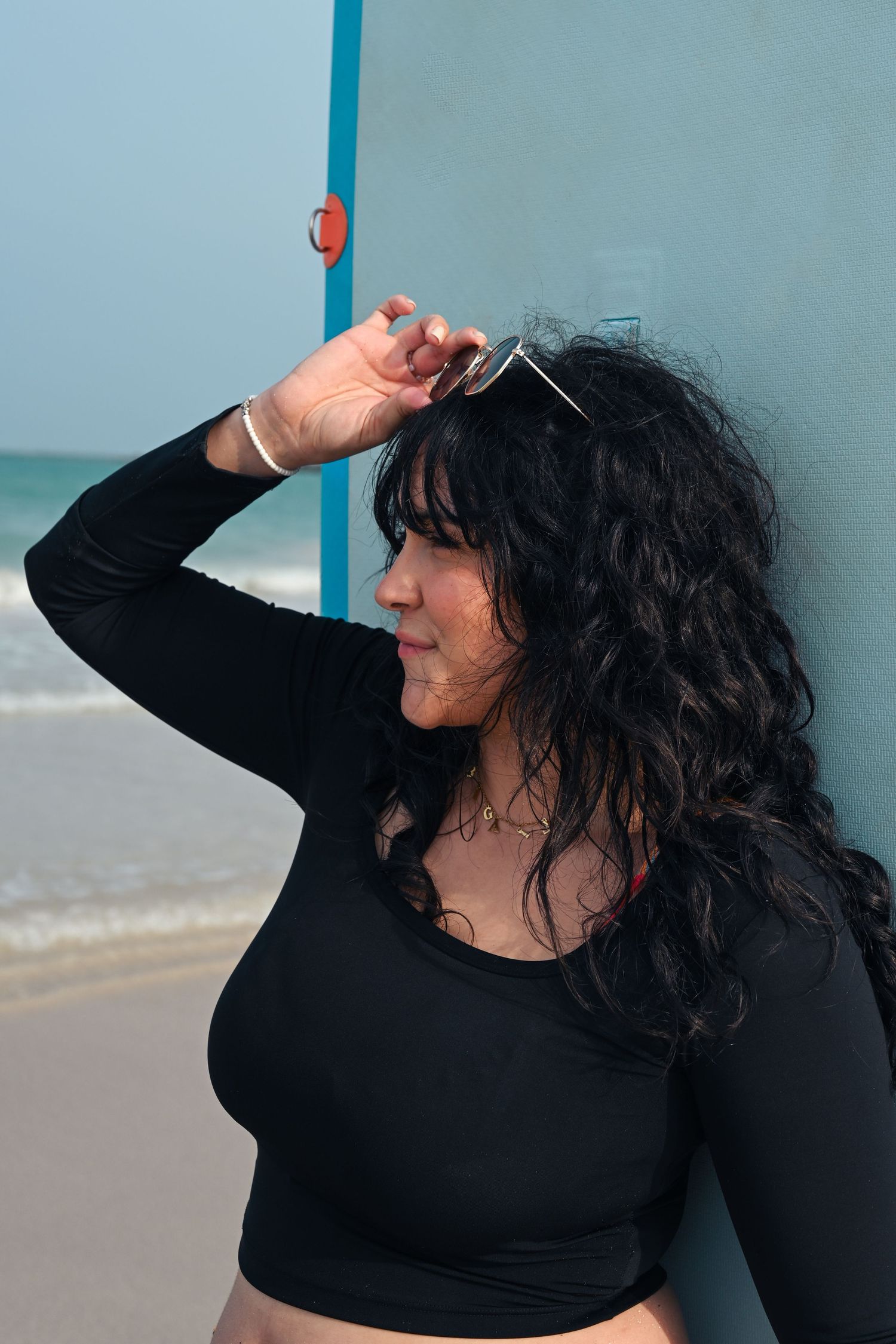 Person in black attire poses against light blue wall near the ocean.