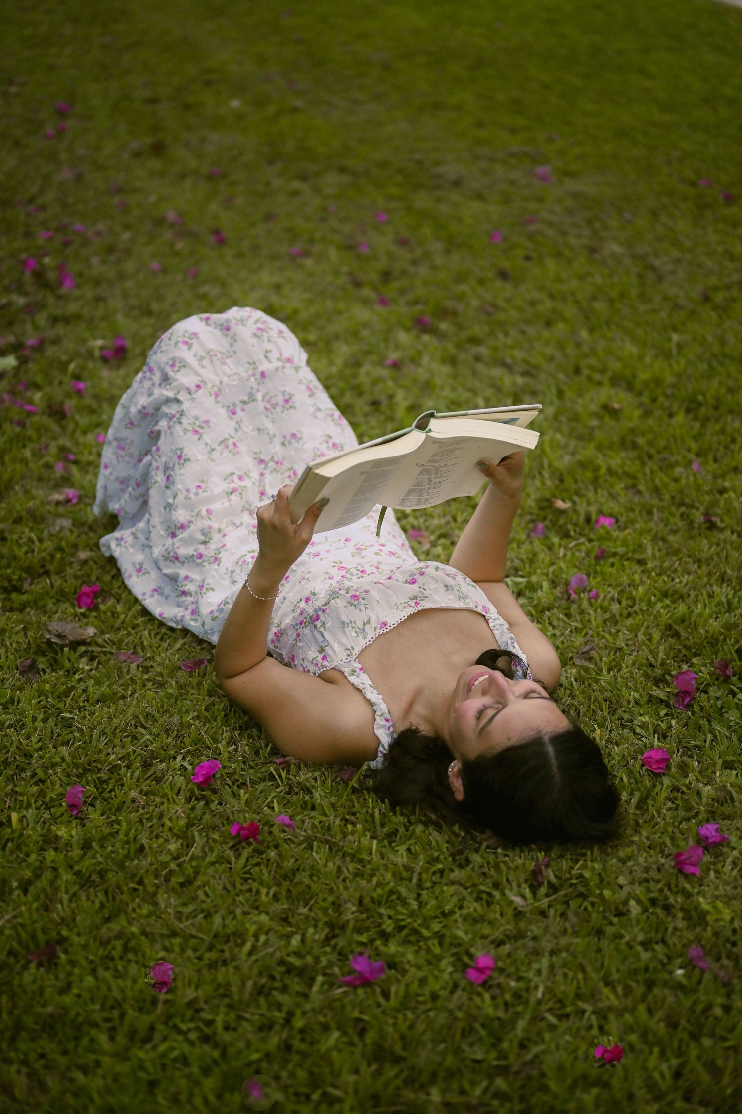 Someone in a white floral dress lies on grass reading among scattered purple flowers.