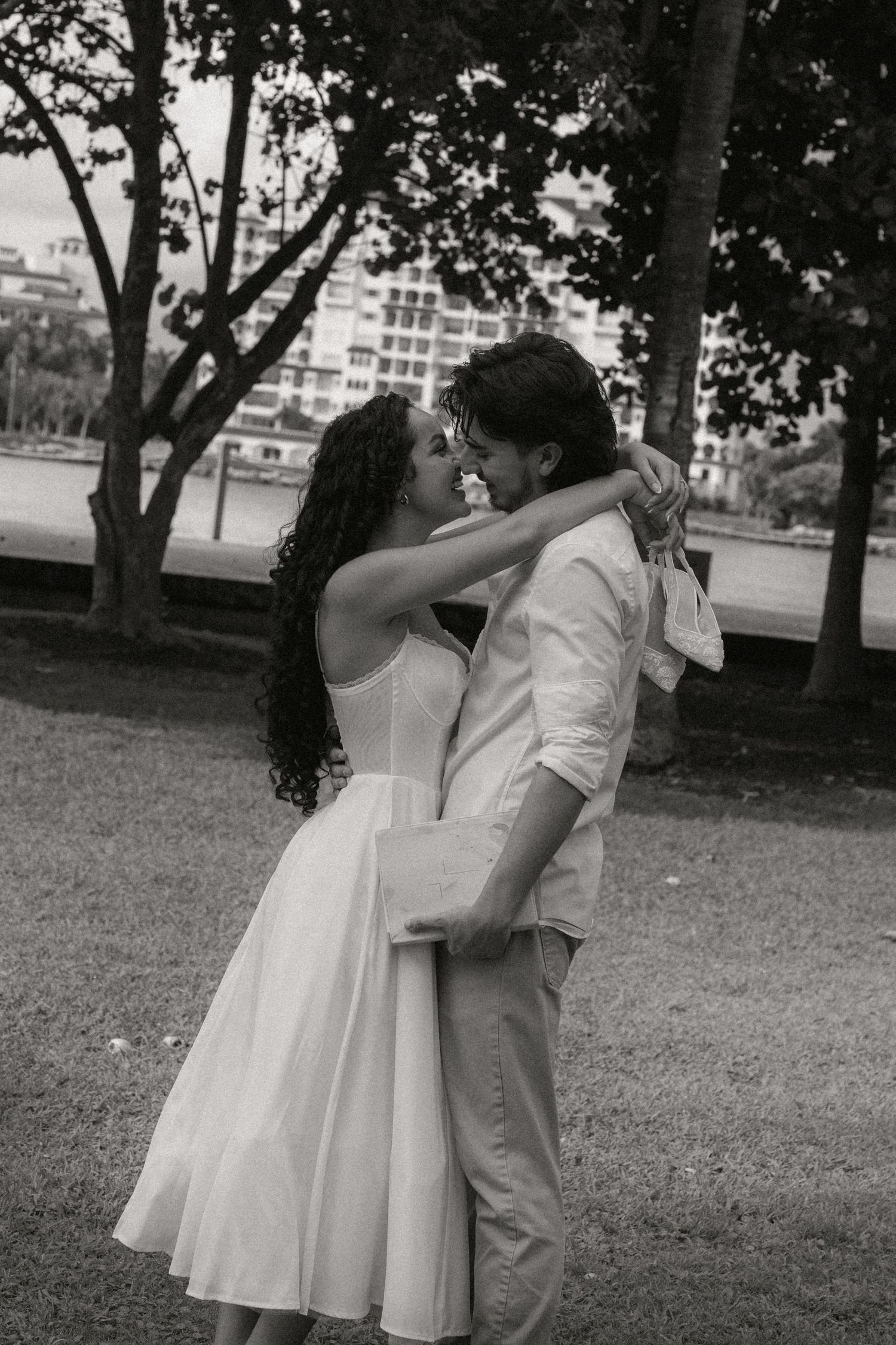 Black and white photo of a couple in white attire sharing a romantic moment in a lush park setting.