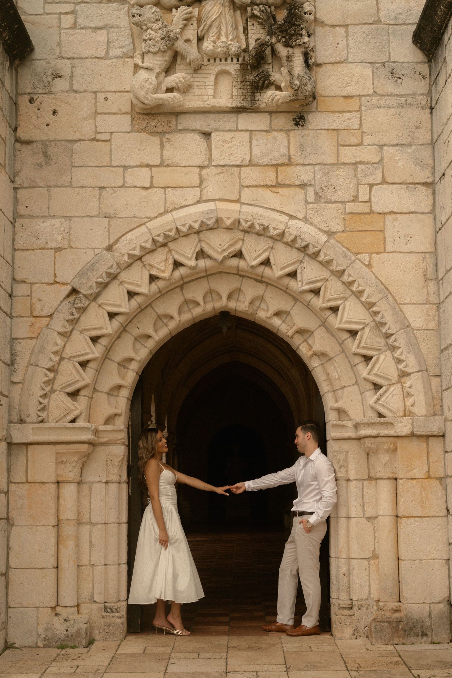 Couple in a white dress and light suit hold hands beneath an ornate stone archway of a historic building during an engagement photo session.