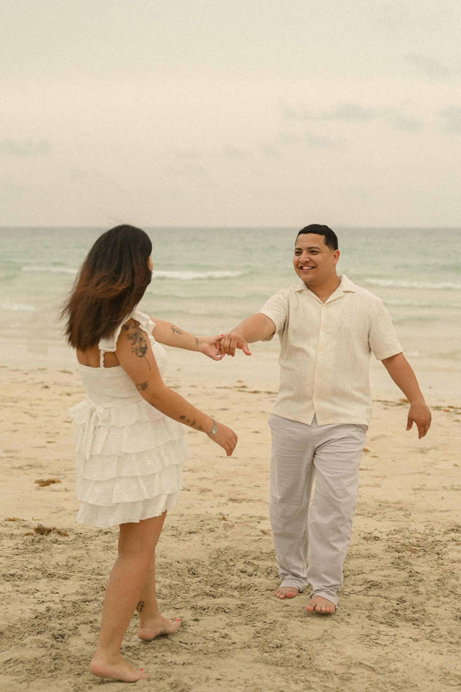 A couple plays and dances together on a sandy beach in matching white outfits.