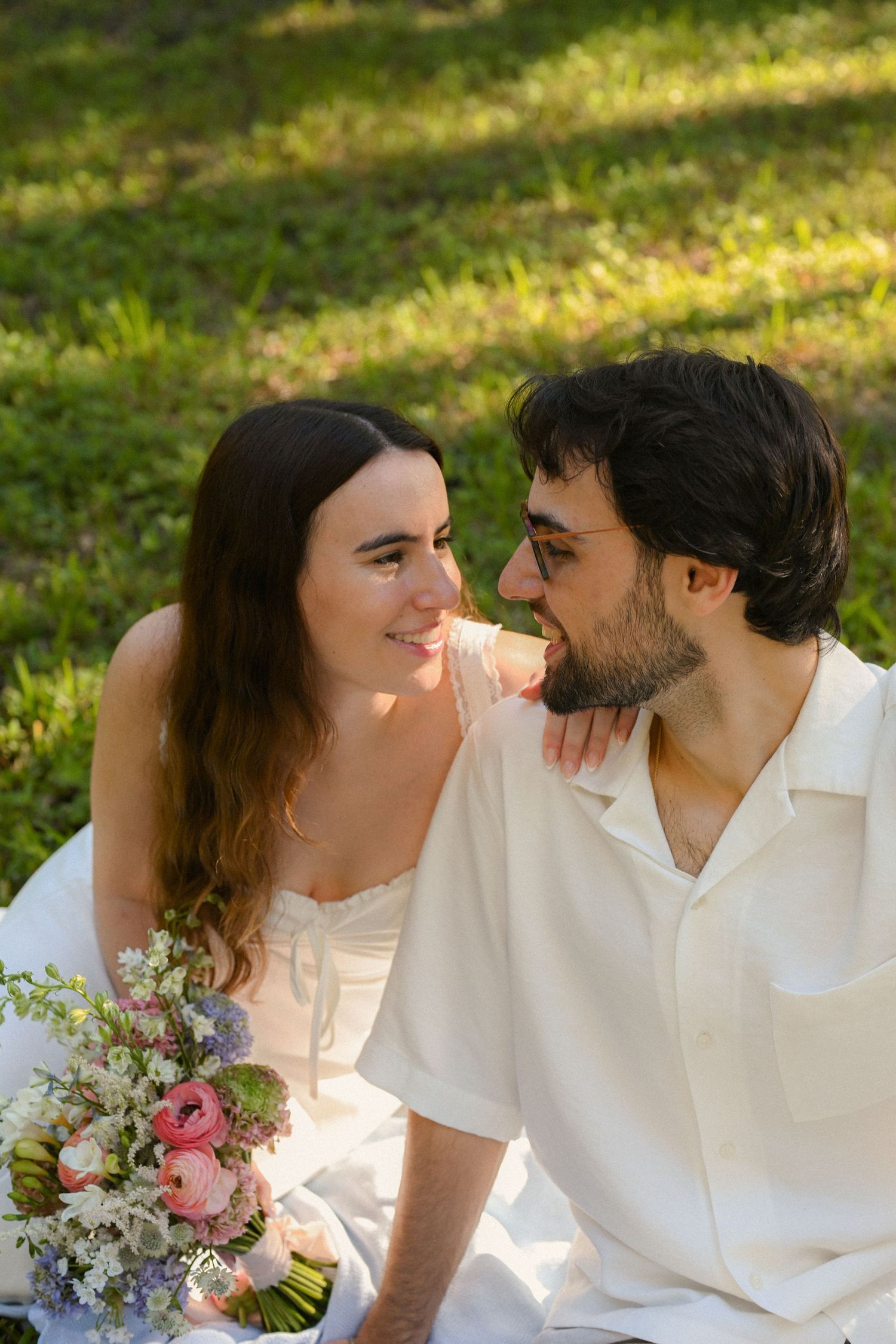 A romantic couple in white outfits share tender moments outdoors while holding a bouquet of pink and purple flowers.