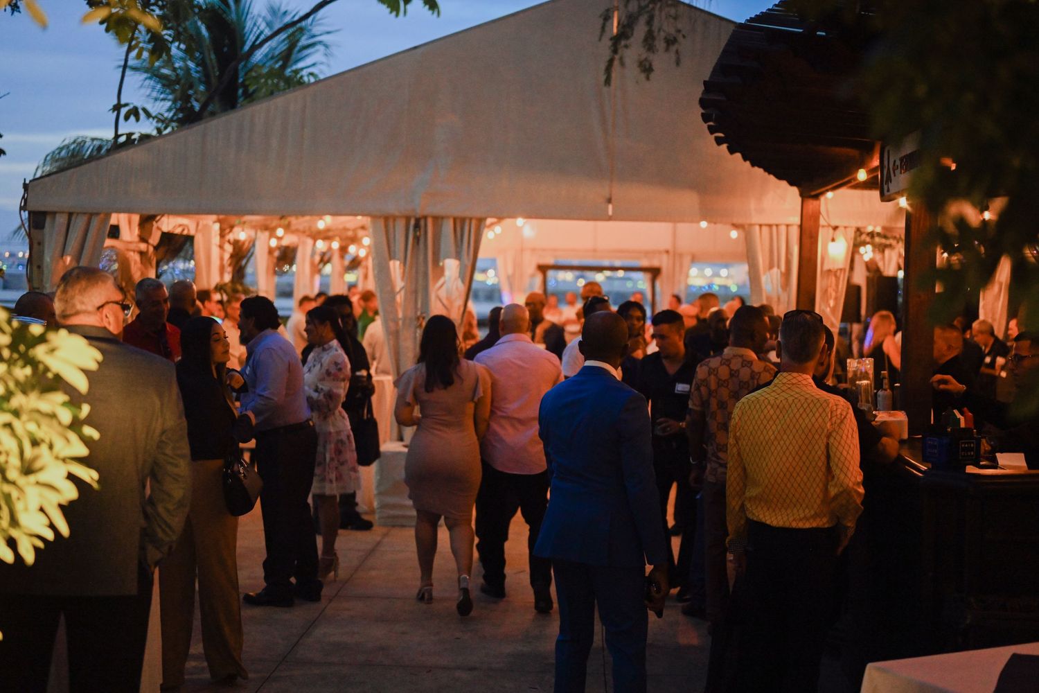 Guests mingle outside a white tent venue at an evening outdoor event with warm string lights illuminating the gathering.