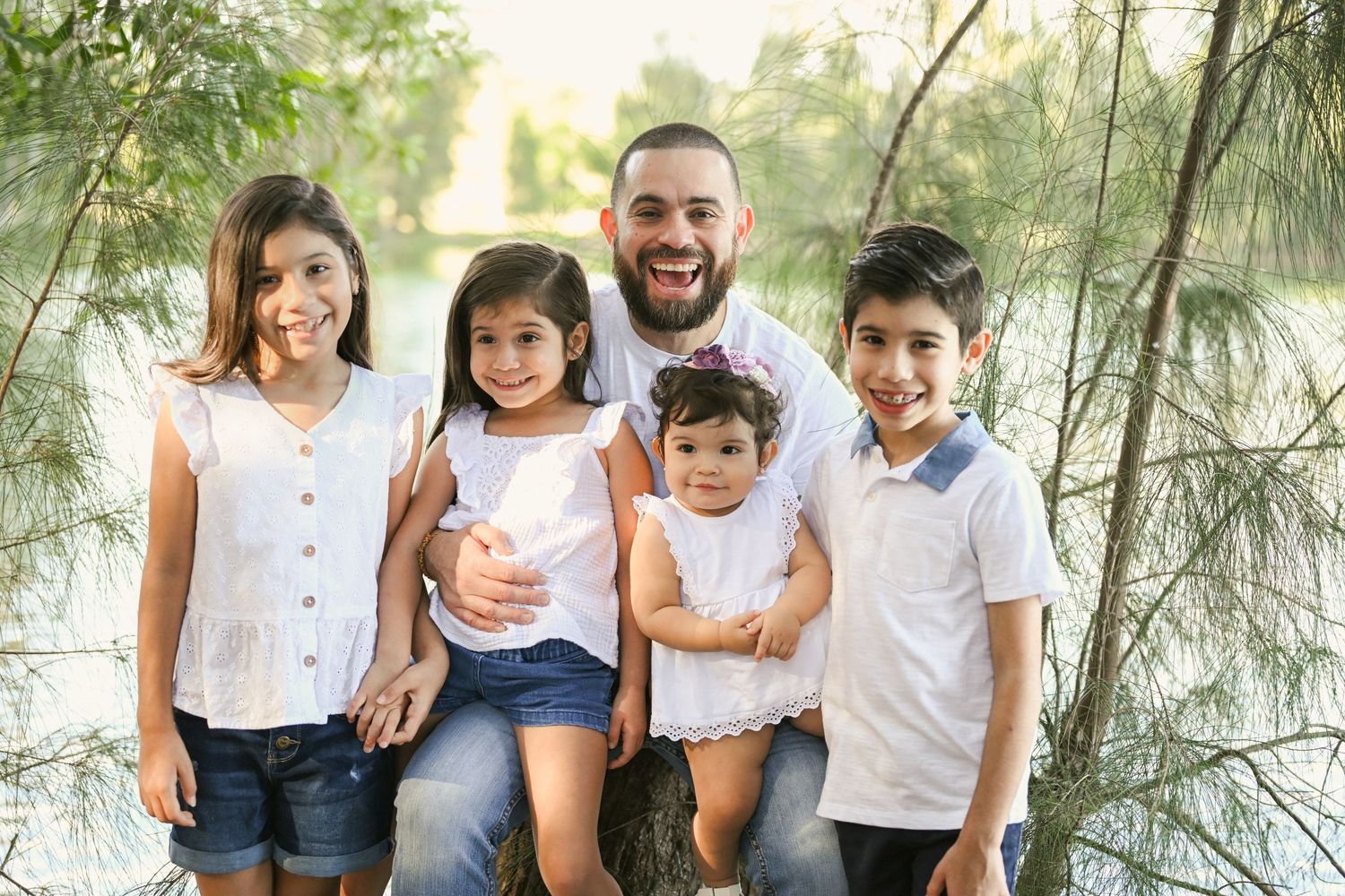 A family wearing coordinated white tops and jeans poses together in a sunlit outdoor setting.