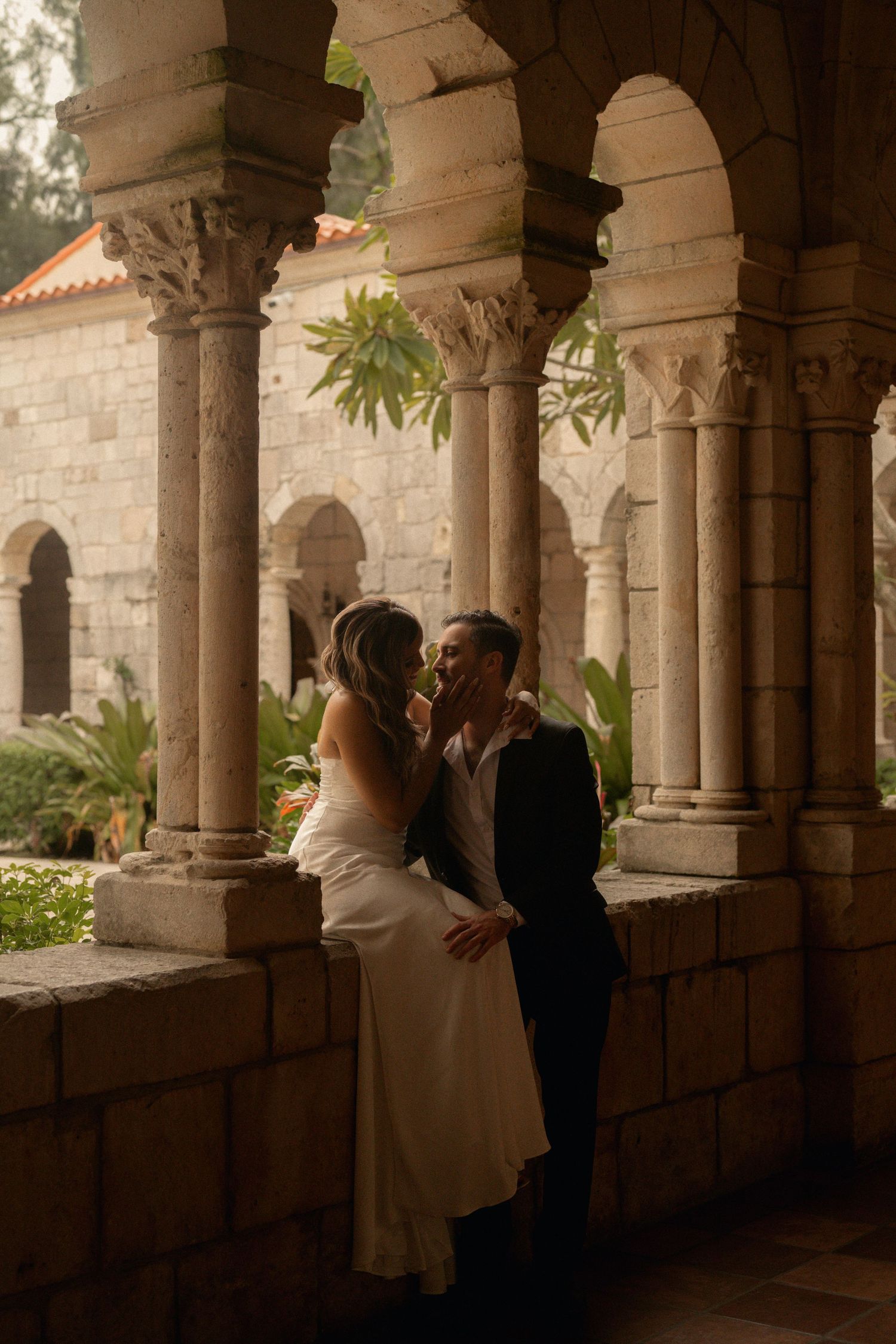 Bride in a white gown and groom in a dark suit share an intimate moment between stone columns at a historic monastery cloister.