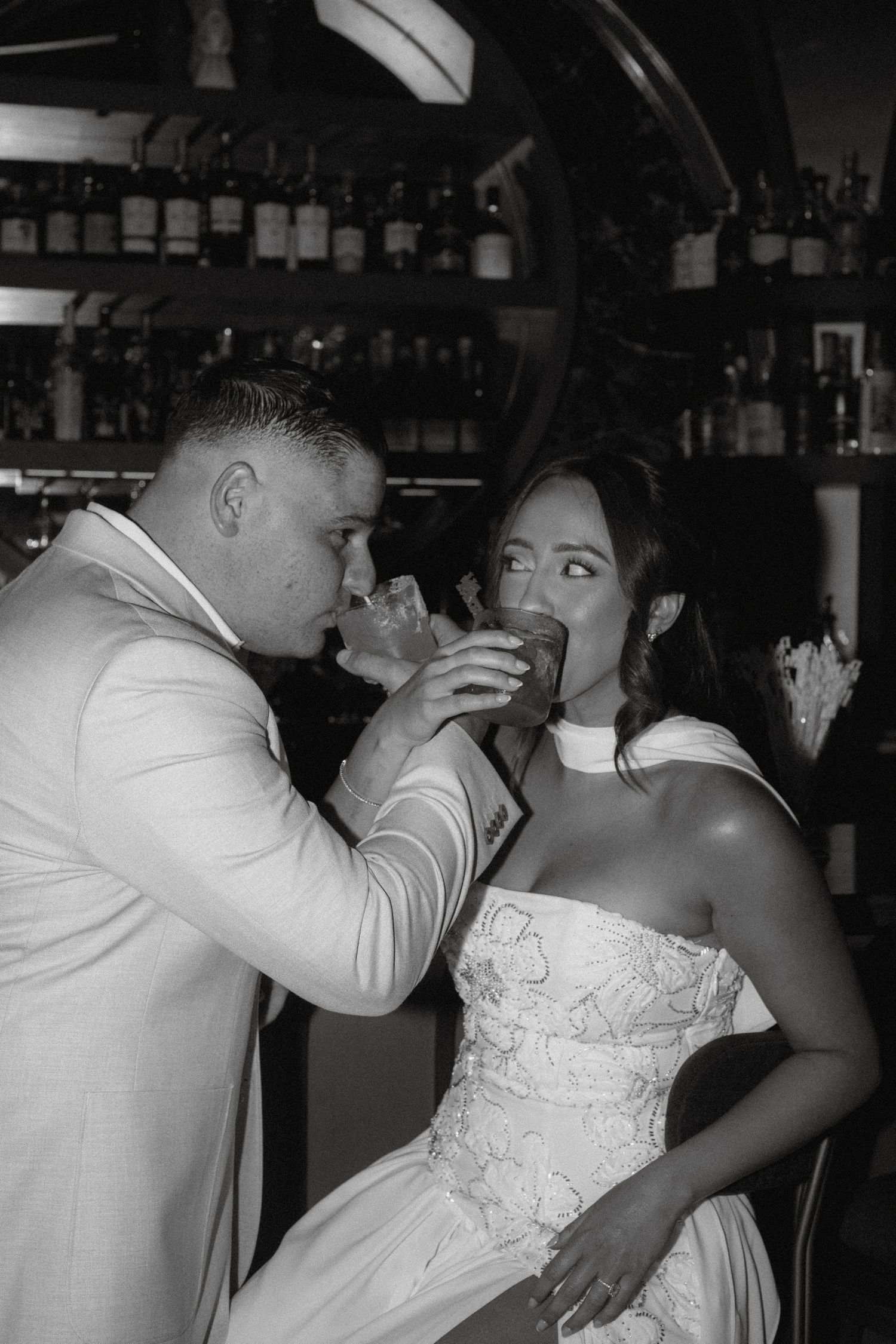 Black and white photo of bride and groom drinking cocktails together at a bar on their wedding day.