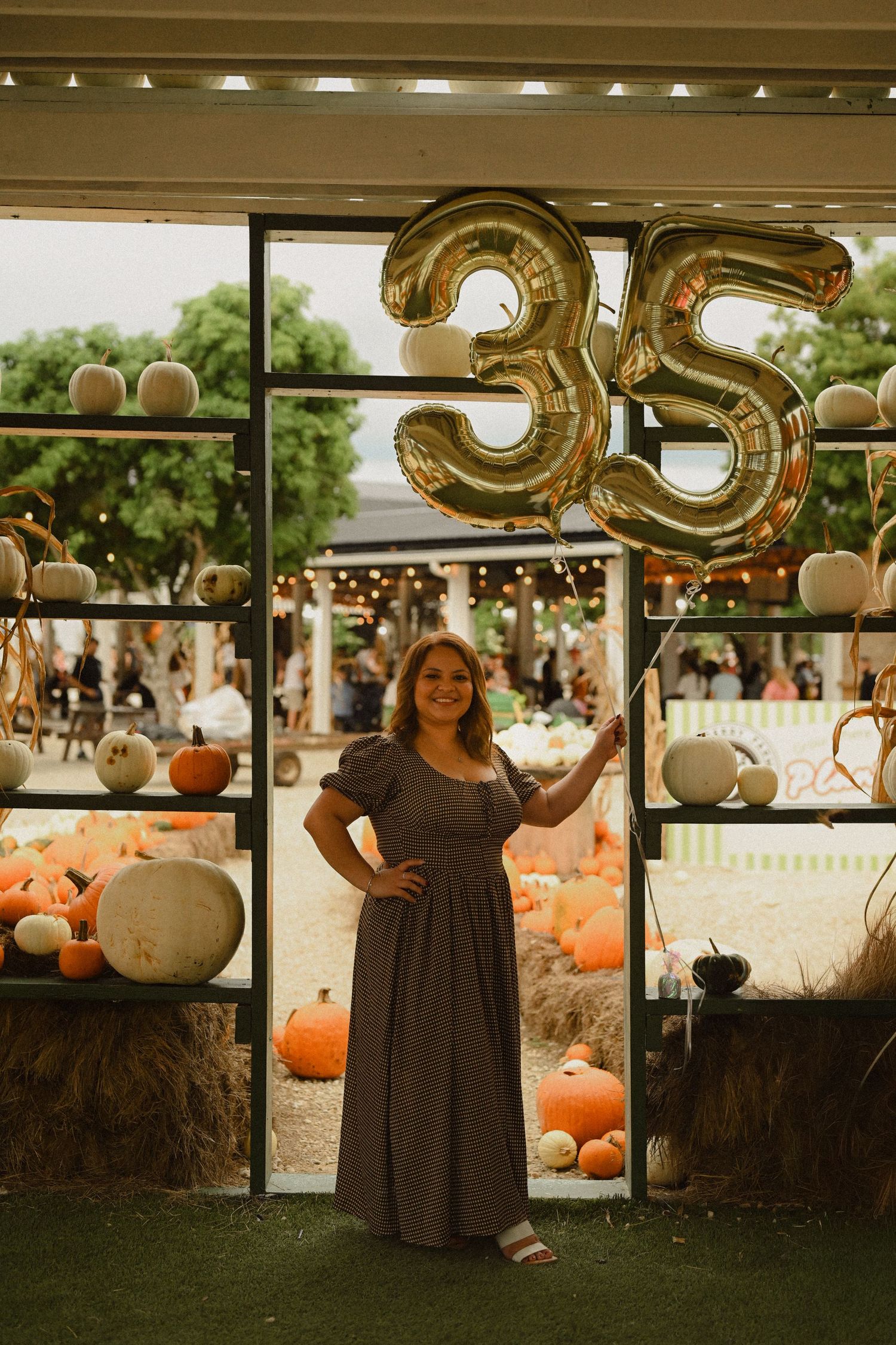 A person in a dark dress stands among pumpkins and gold number balloons at an outdoor autumn celebration.