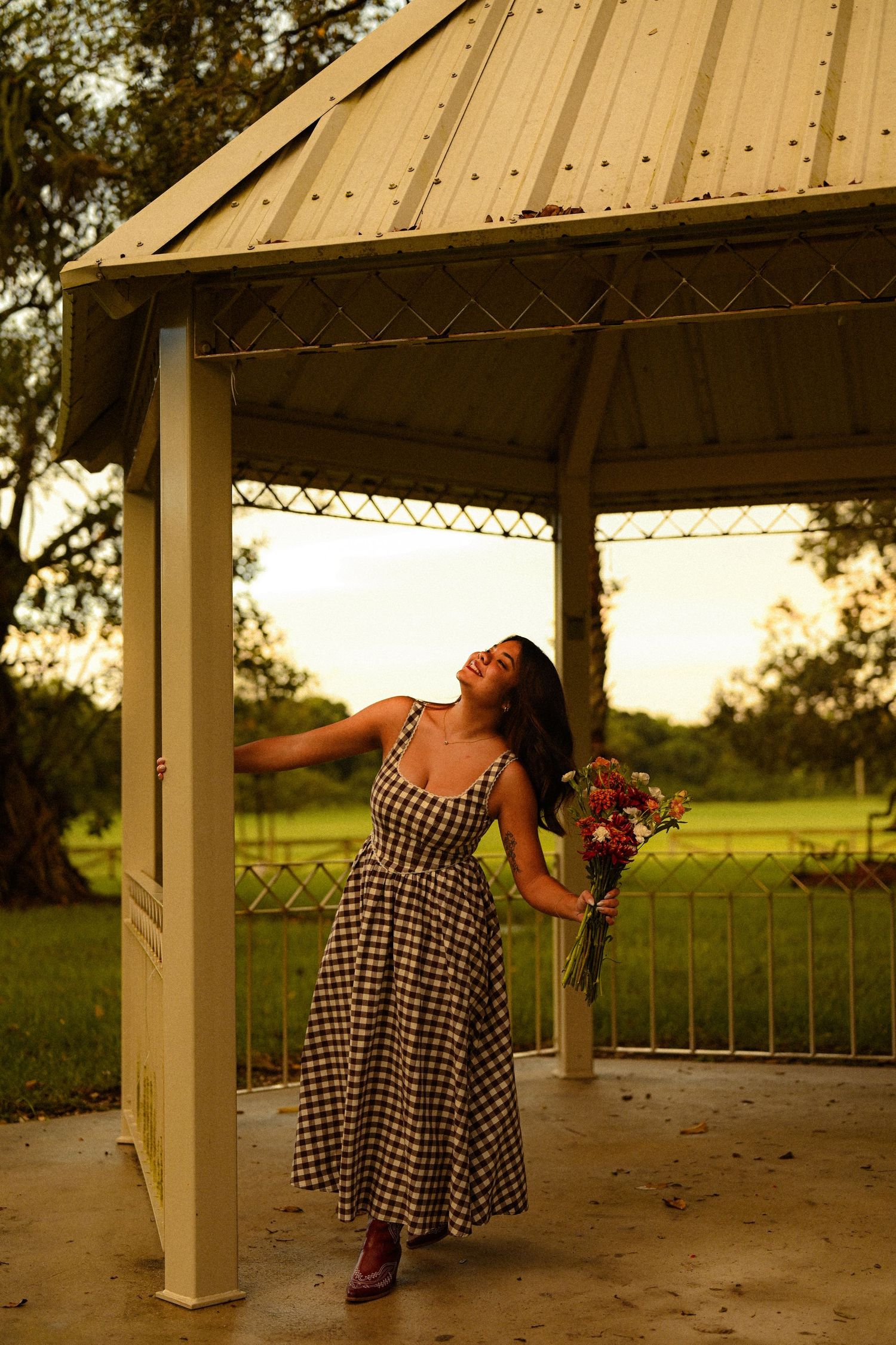 Figure in gingham dress poses dramatically in a white gazebo at sunset.