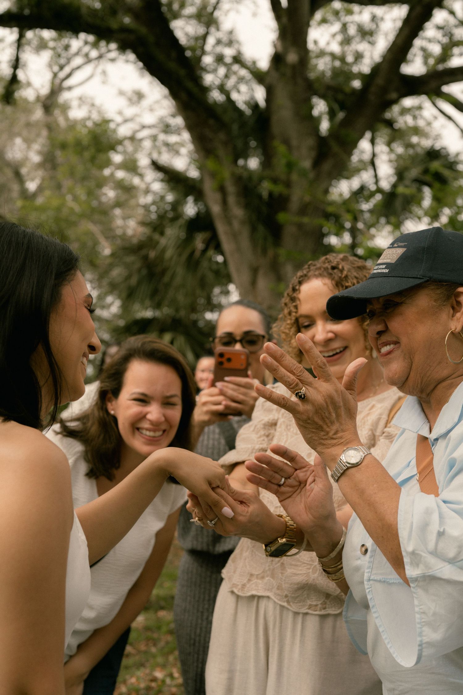 Joyful group of friends and family admire an engagement ring outdoors under a large tree.