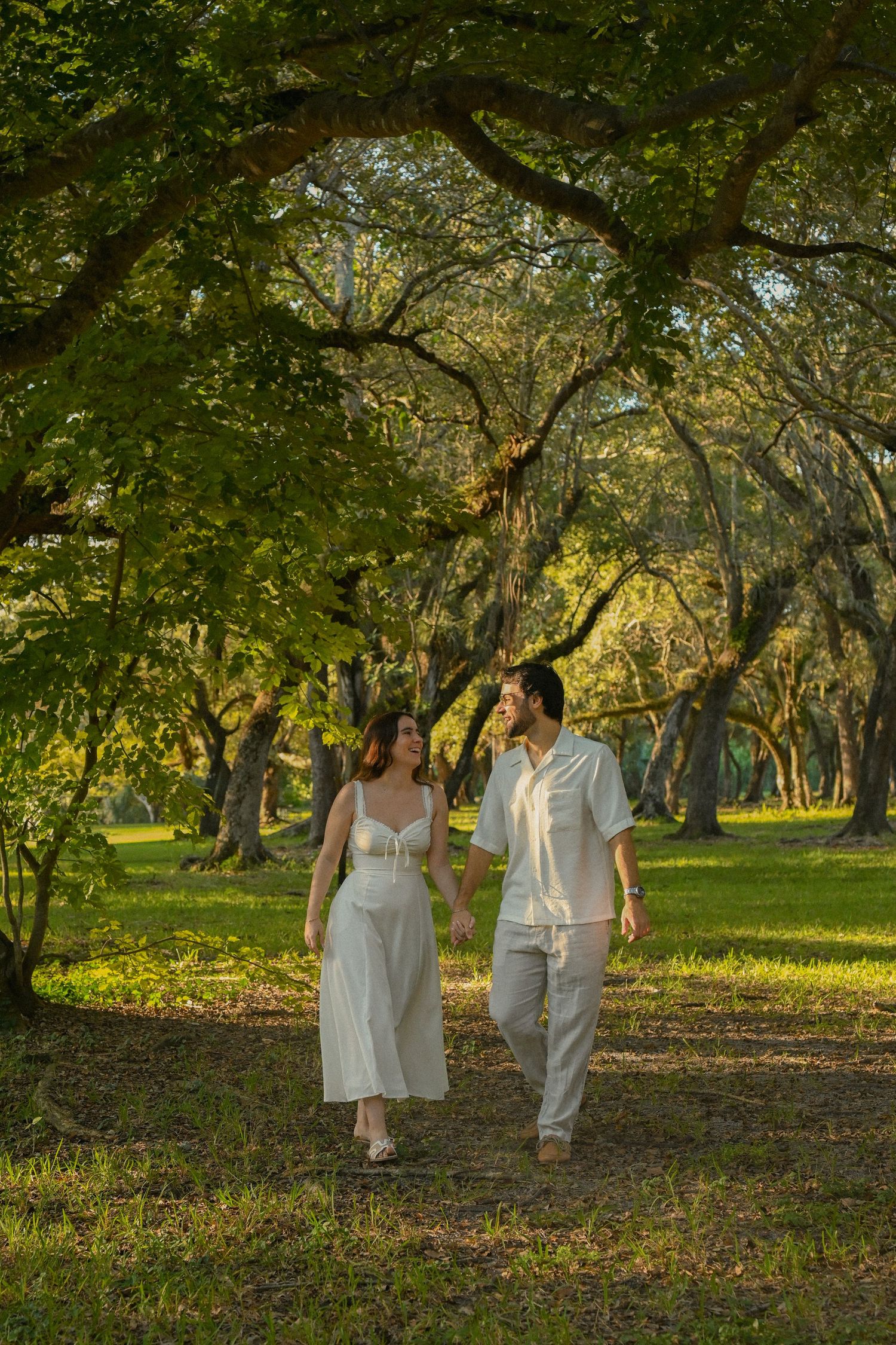 Couple in white attire walks hand in hand through a sunlit garden path under lush green trees at golden hour.