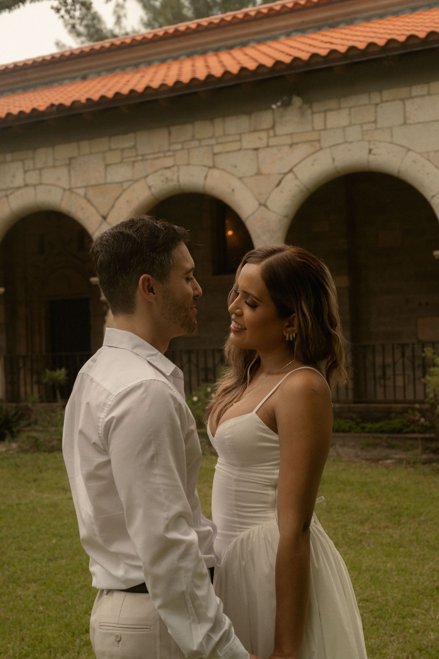 Smiling couple standing face to face on a lawn in front of a rustic stone building with arches and a terracotta roof.