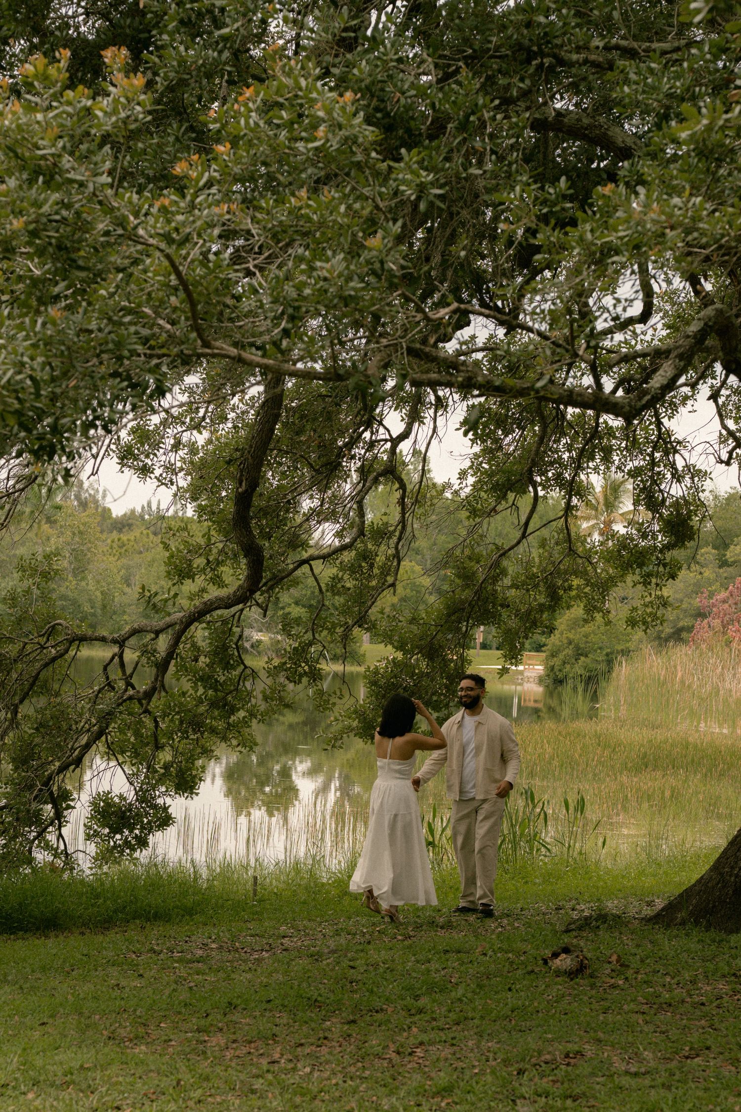 Couple holding hands under a large oak tree by a serene lake, dressed in elegant white and beige wedding attire.