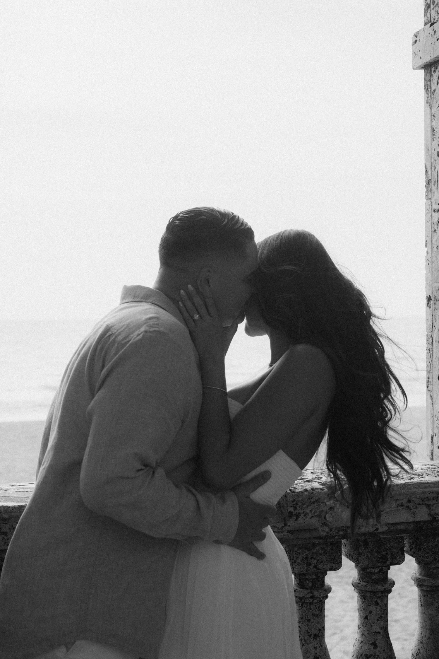 Black and white photo of a romantic couple embracing and kissing on a seaside stone balcony.