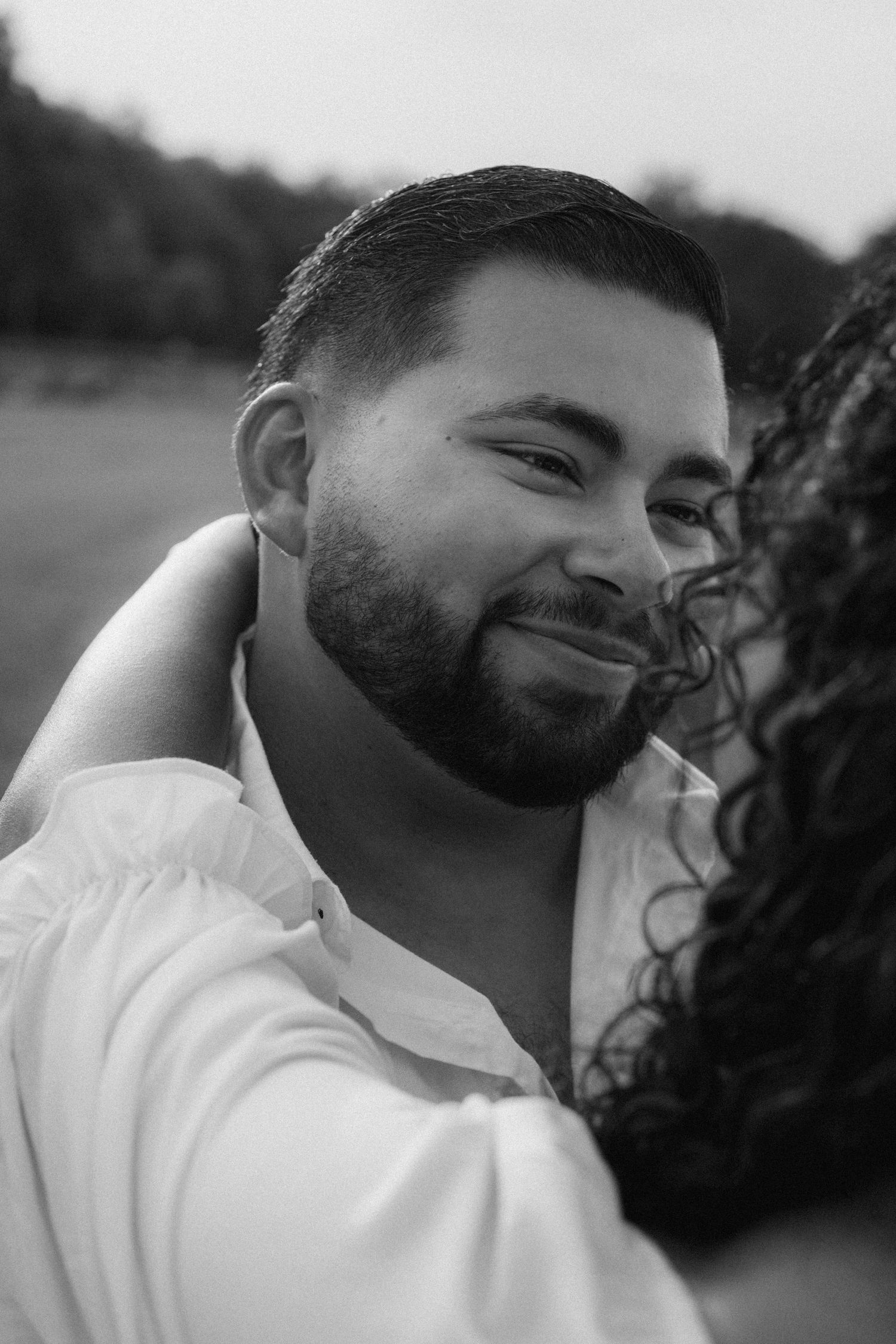 Black and white portrait of a smiling man in a white shirt with a woman partially visible beside him outdoors.