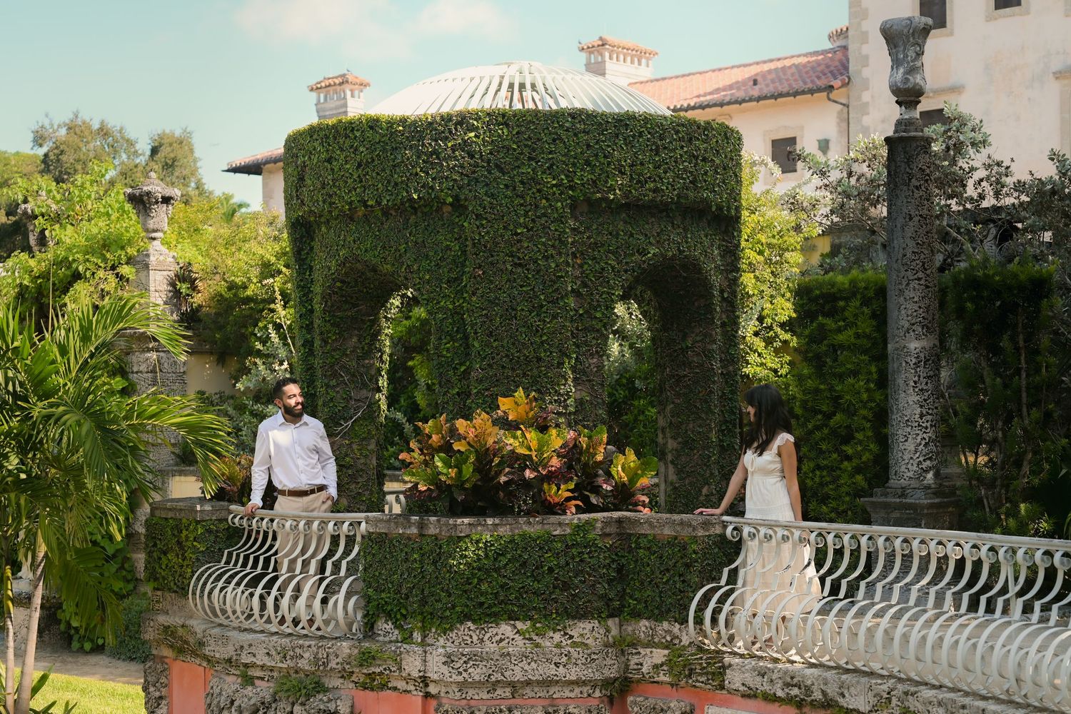 A decorative stone balcony with ornate railings overlooks a lush garden with palm trees and flowering plants.