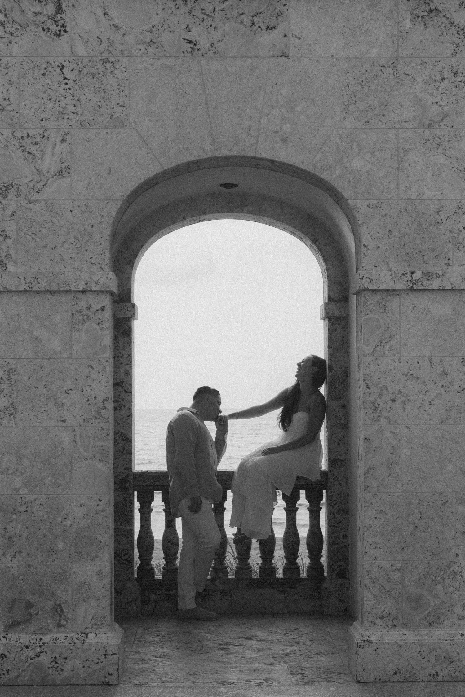 Black and white photo of a couple in an arched stone doorway, woman sitting on balcony railing touching mans face by the sea.