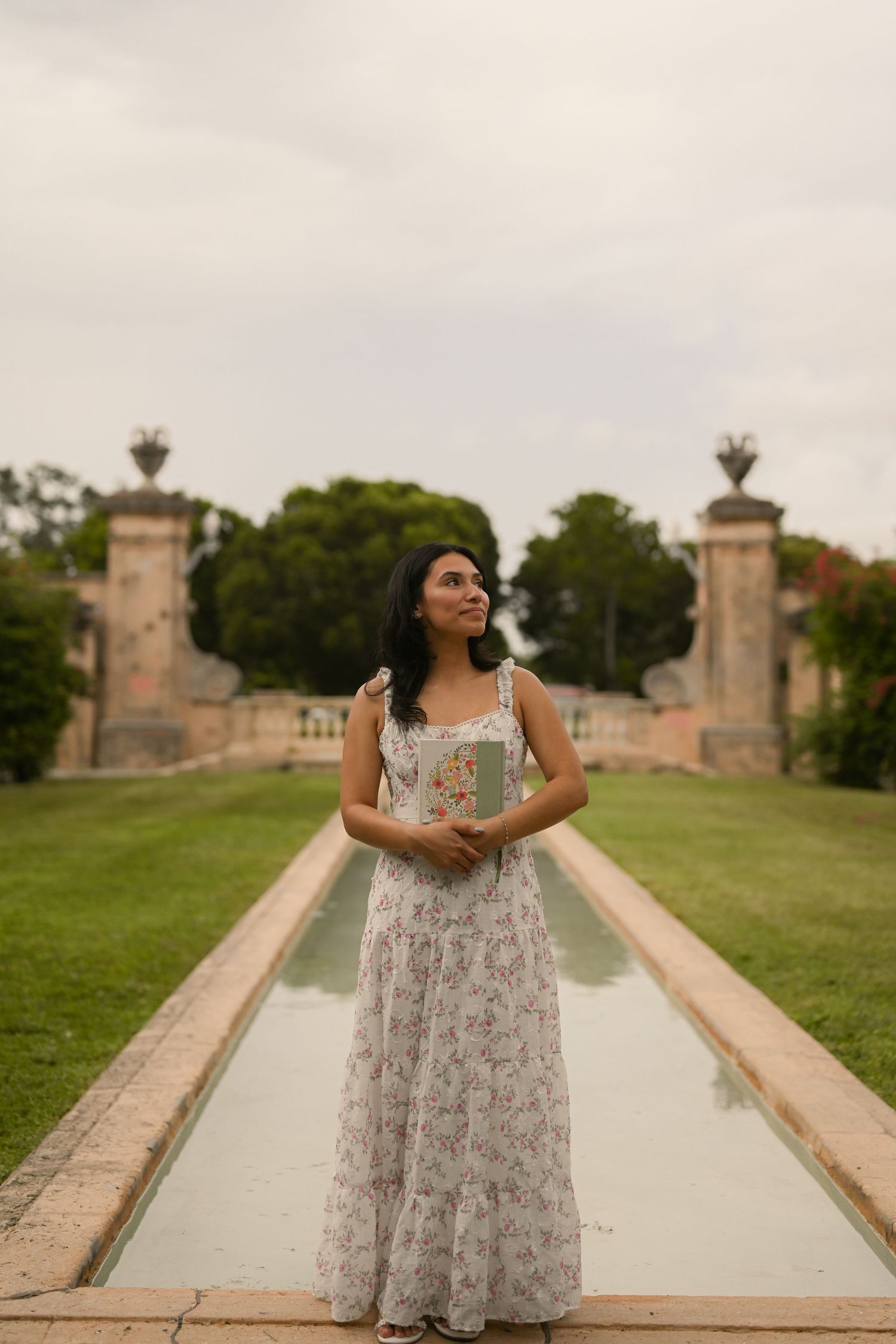 A person in a flowing dress stands along a reflecting pool surrounded by classical architecture.