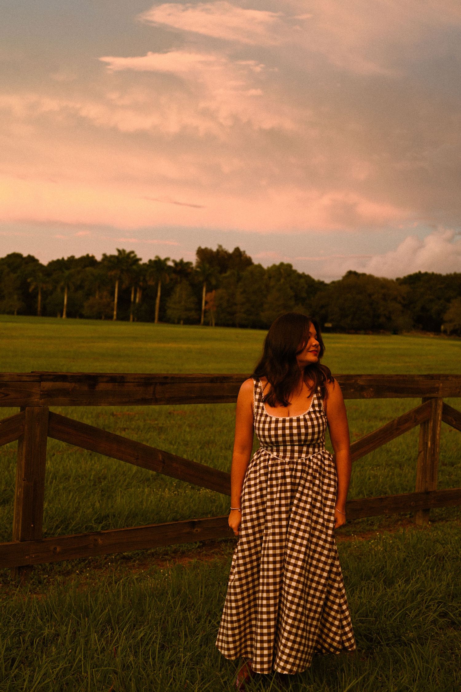 Person in checkered dress stands by wooden fence in field during pink sunset.