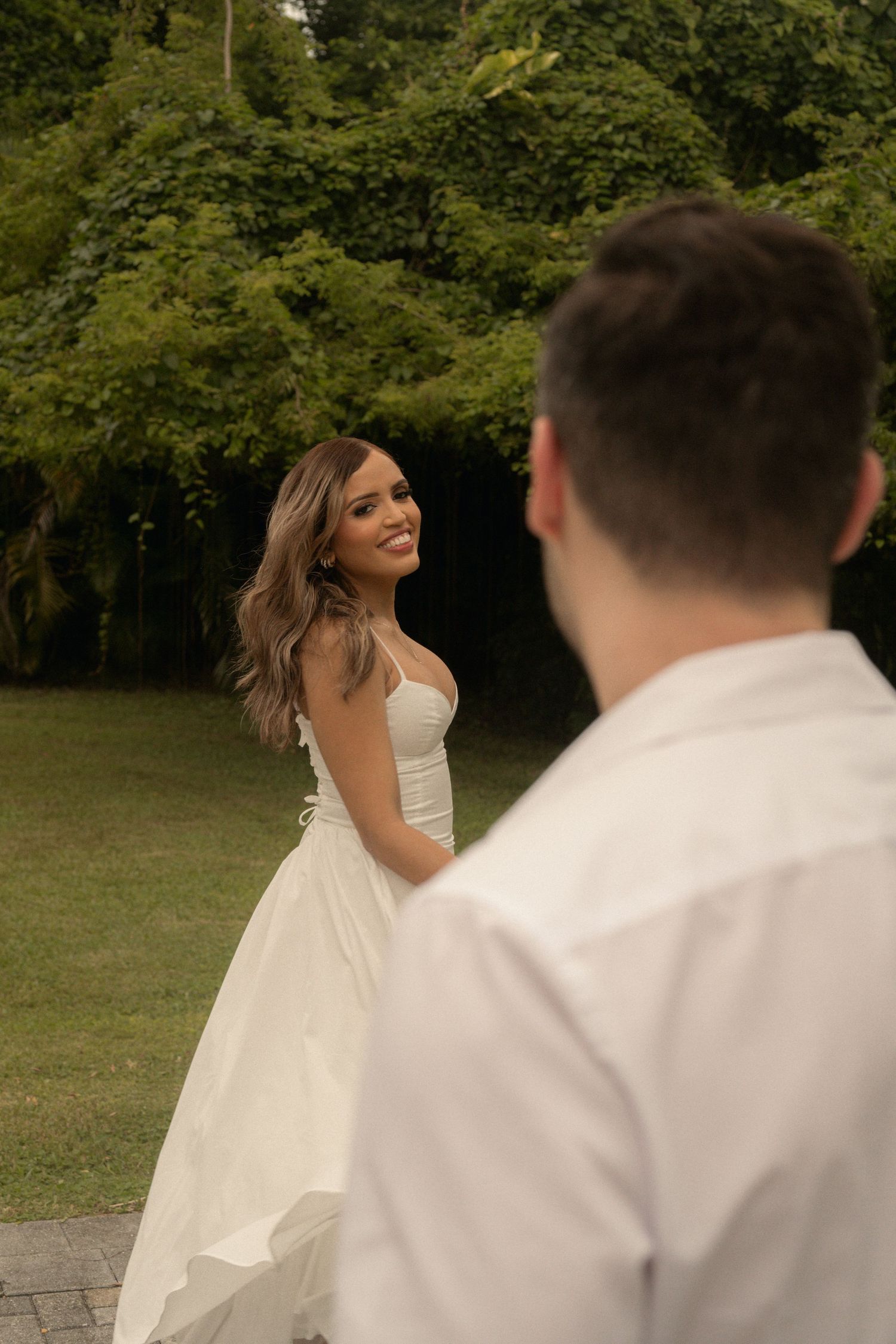Smiling bride in a white strapless wedding gown looks back over her shoulder at a groom in the foreground, with lush green trees behind.