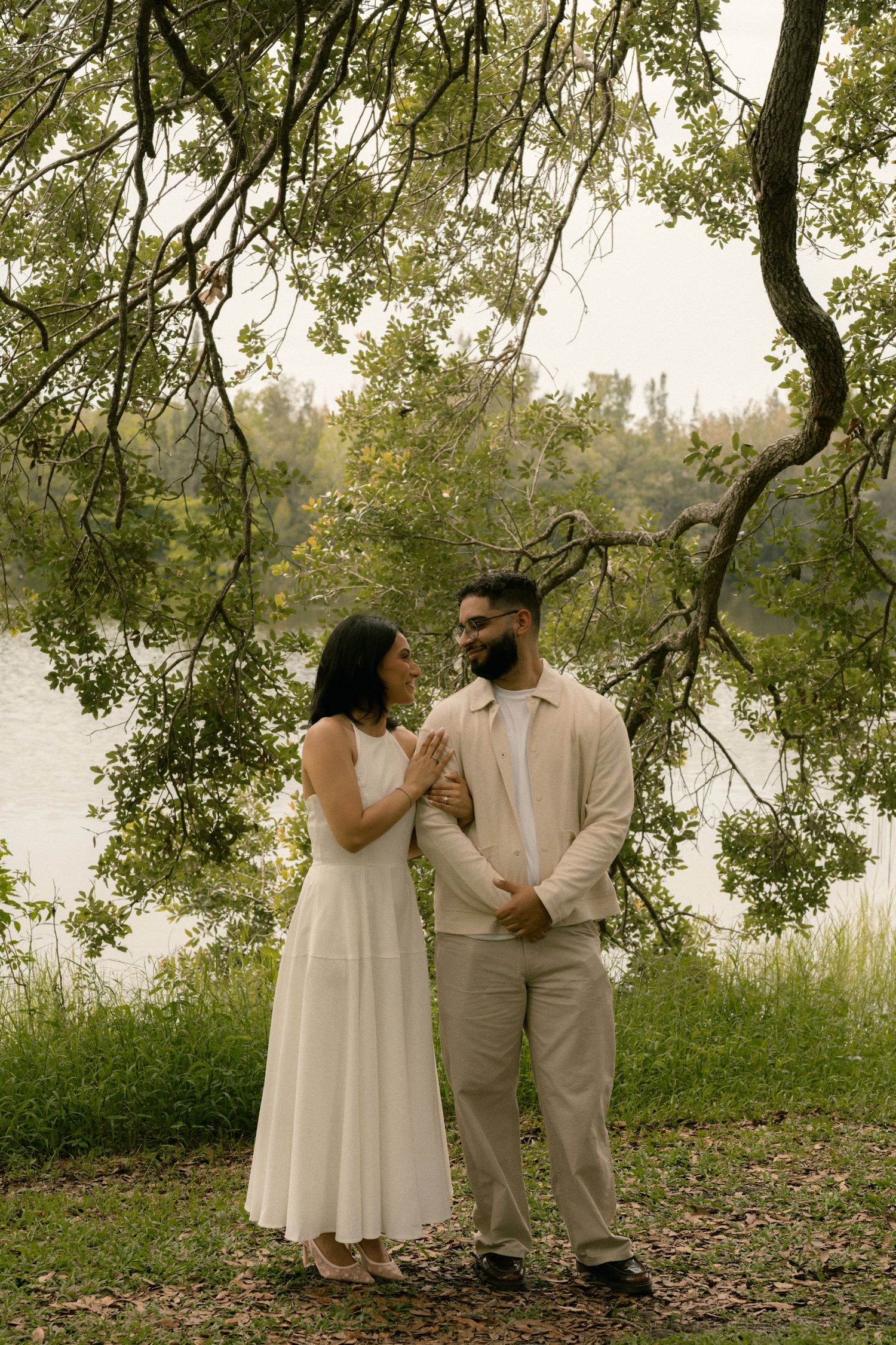 Newlywed couple holds hands and gazes at each other beside a serene lake under trees.