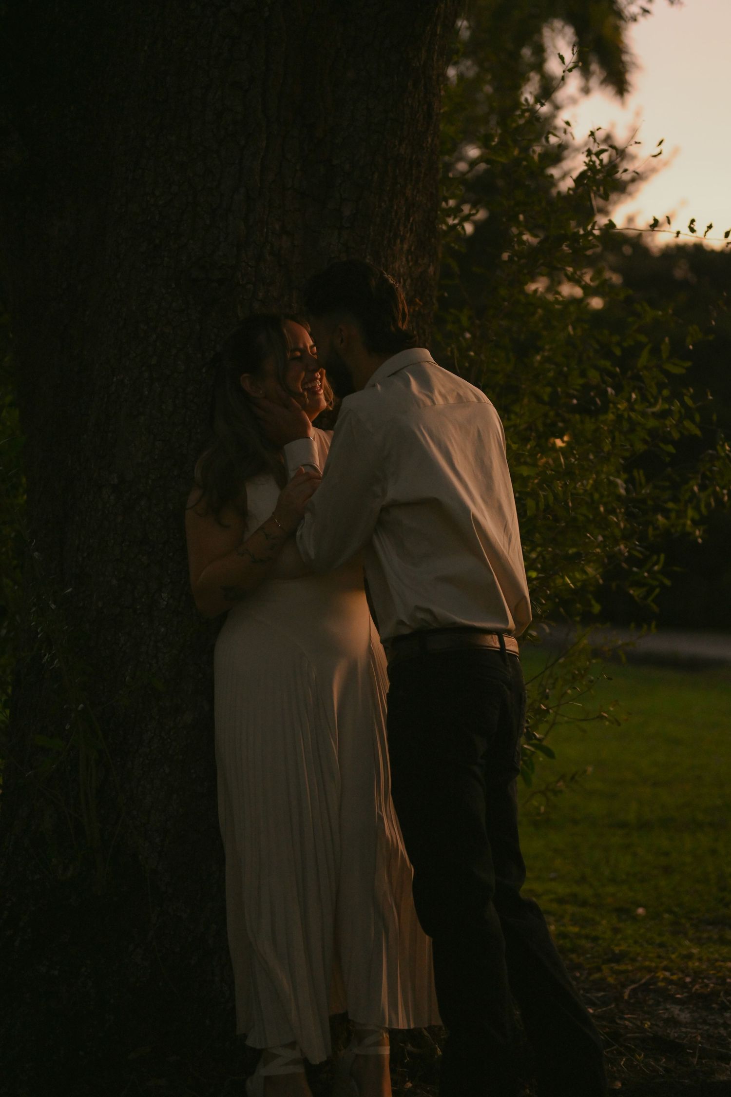 Couple sharing a romantic moment at dusk, leaning against a large tree, woman in white dress, man in white shirt.