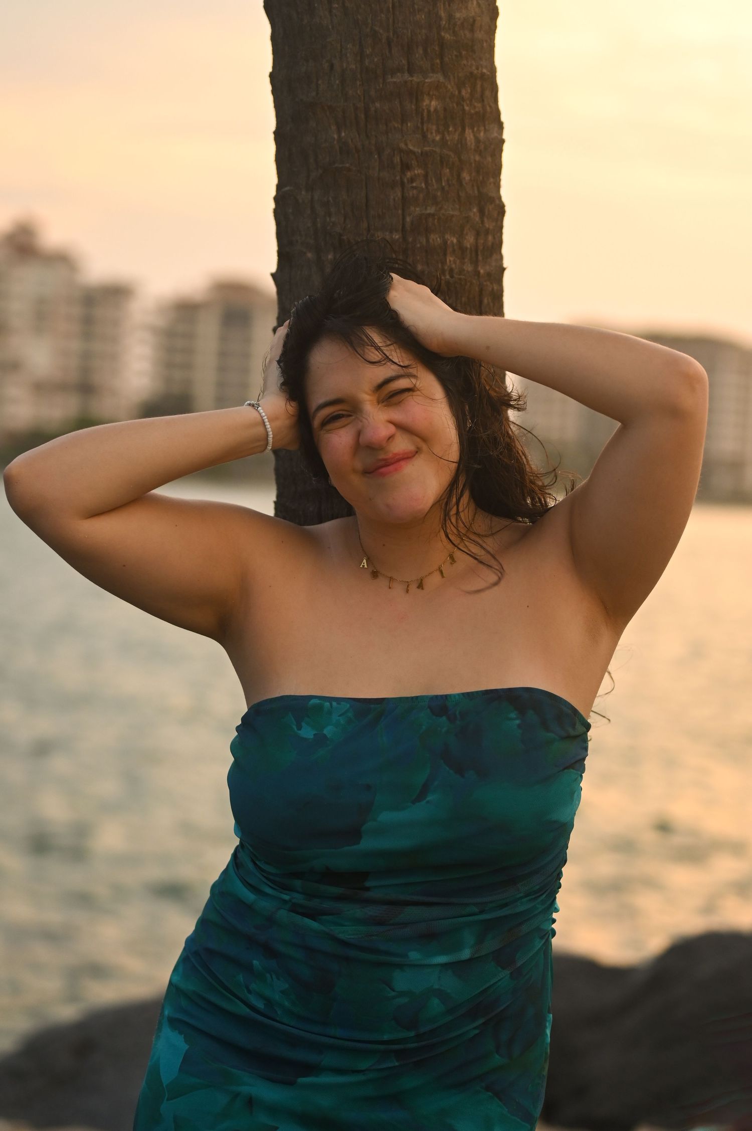 Candid poses in a teal strapless dress against a palm tree at sunset.