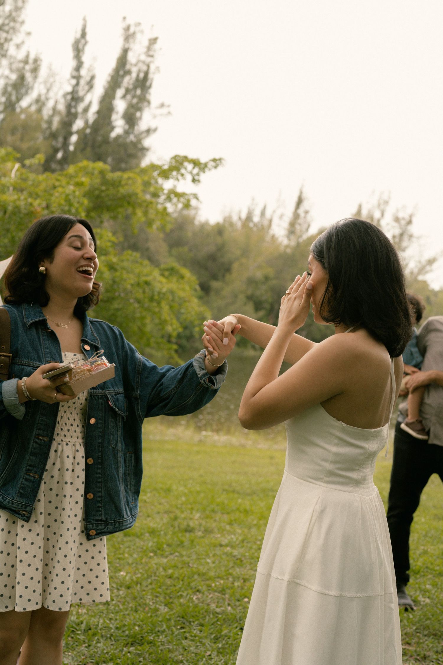 Two women laugh and celebrate outdoors in a lush green park, one wearing a white dress and the other a denim jacket.