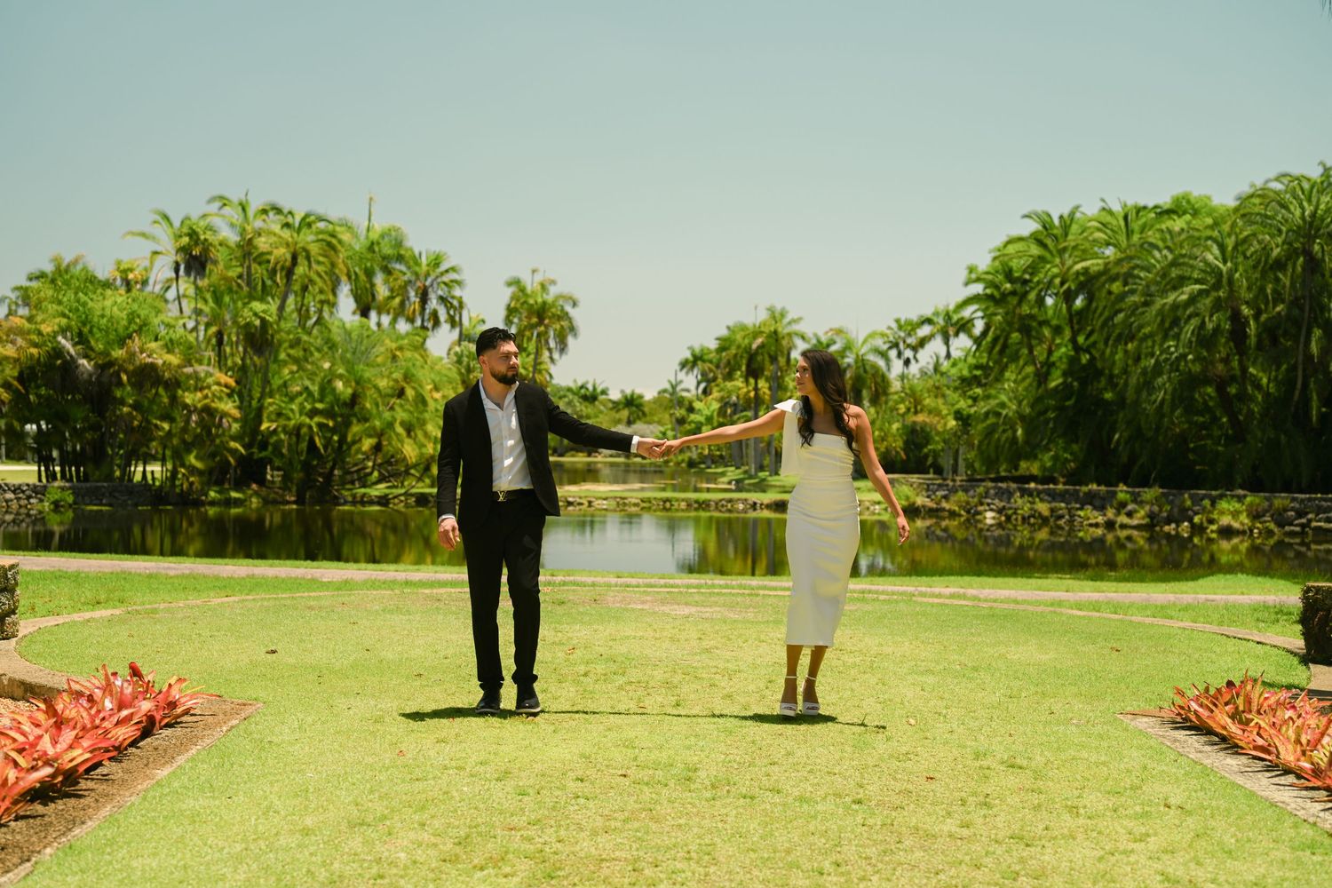 A couple walks together on a lush green lawn with palm trees and tropical landscaping.
