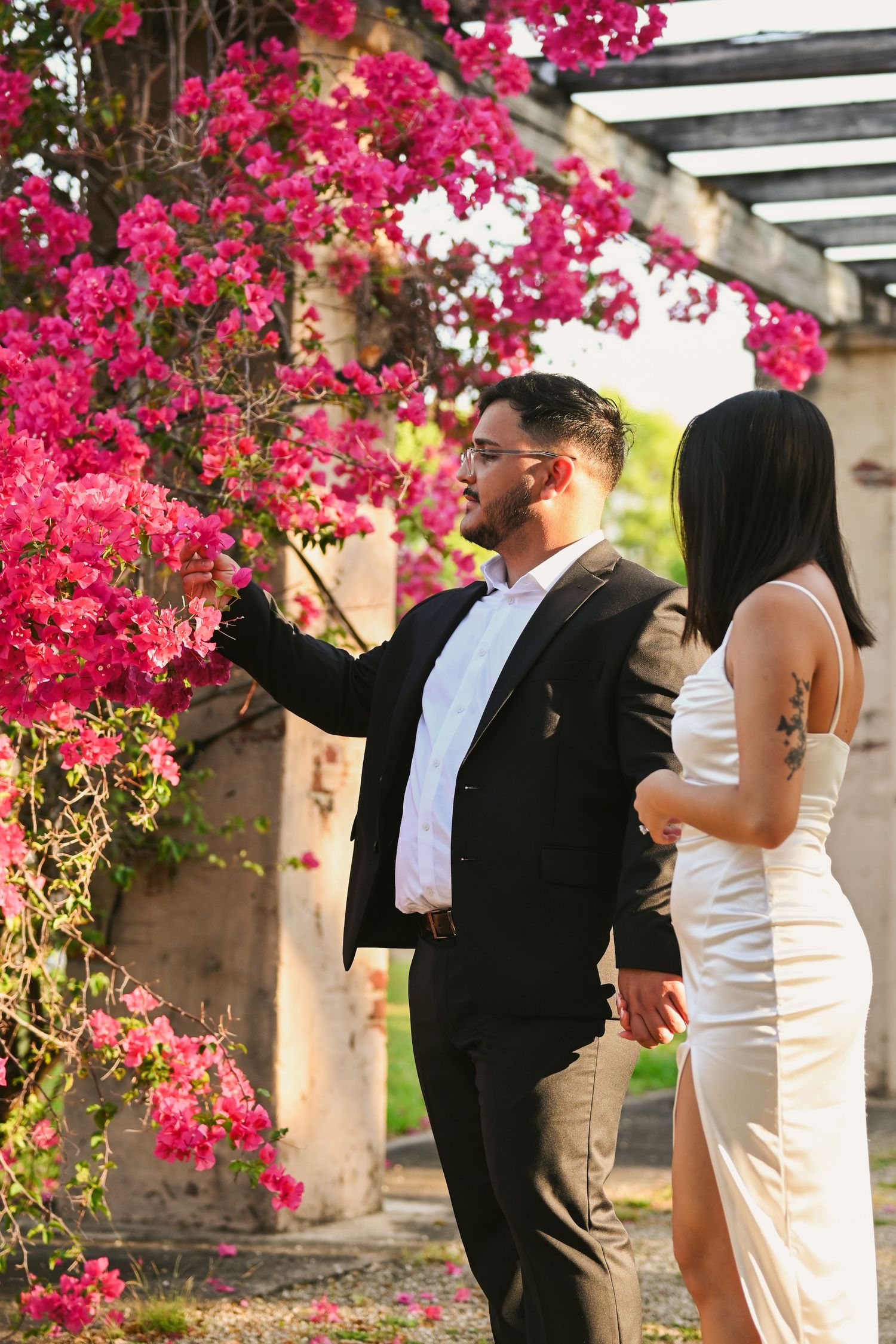 Couple in black and white attire sharing playful moments under a flower-covered pergola.