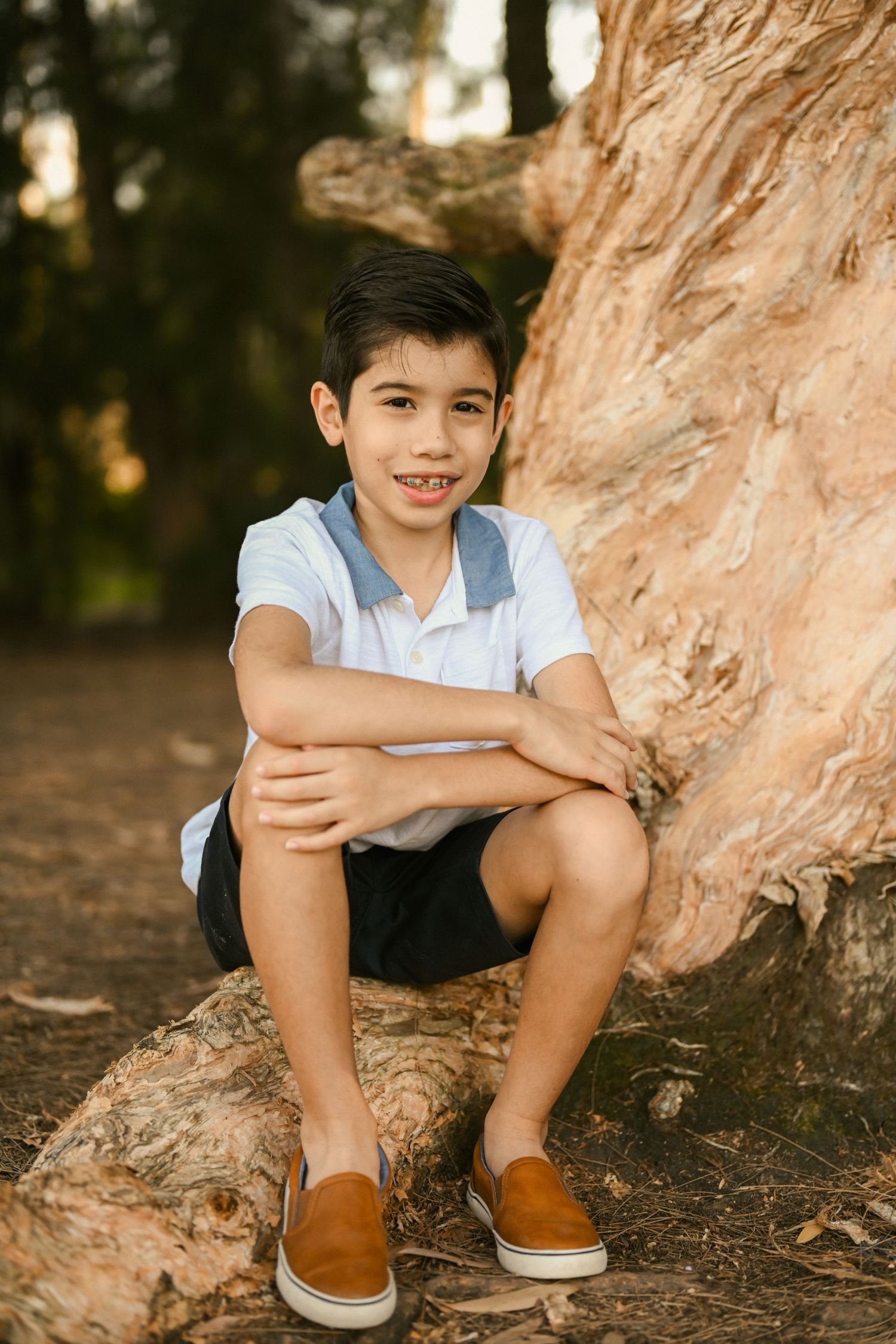Child sits casually on tree roots wearing brown shoes and white shirt during golden hour photography session.