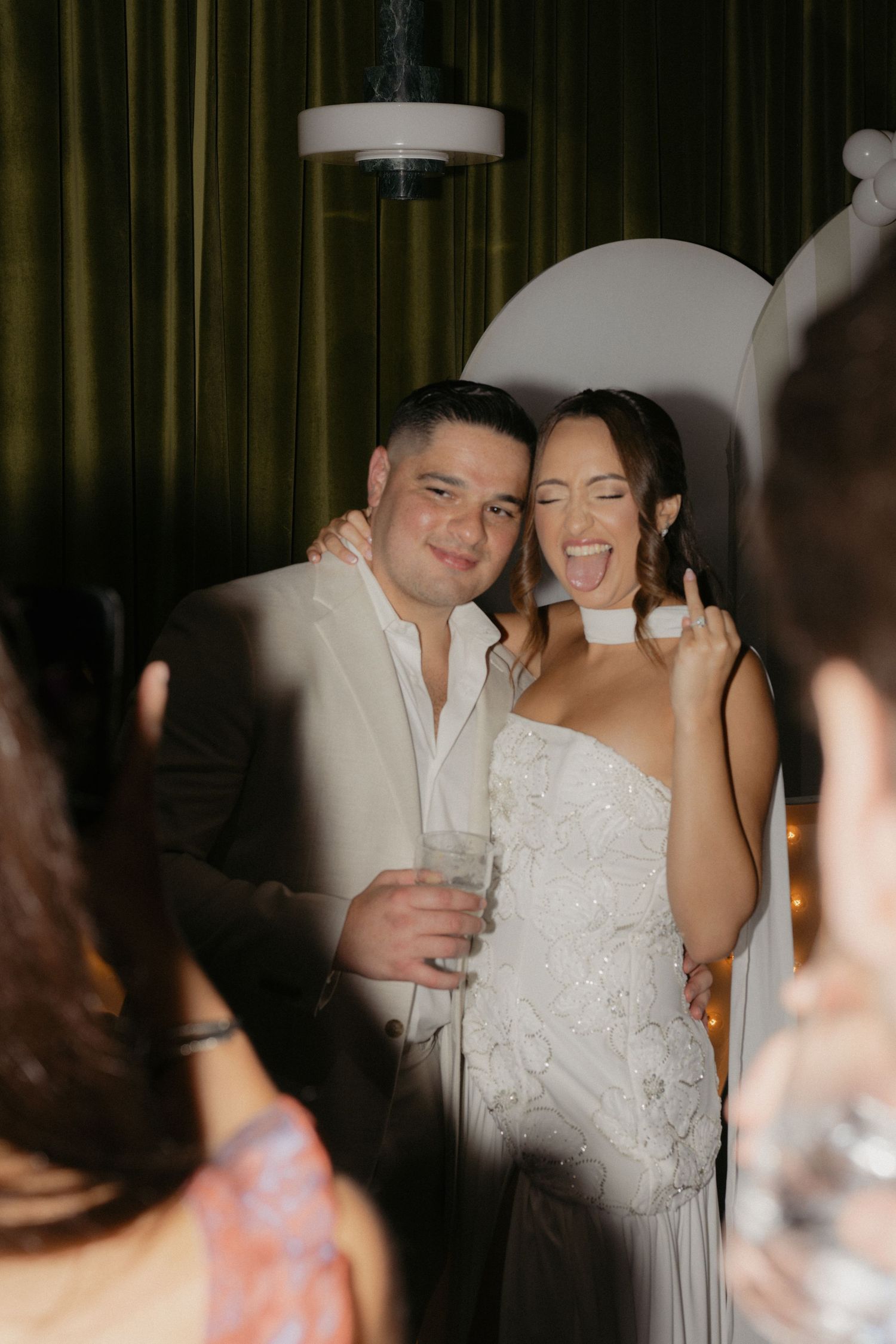 Joyful bride and groom posing at wedding reception, bride making fun gesture near photo booth.