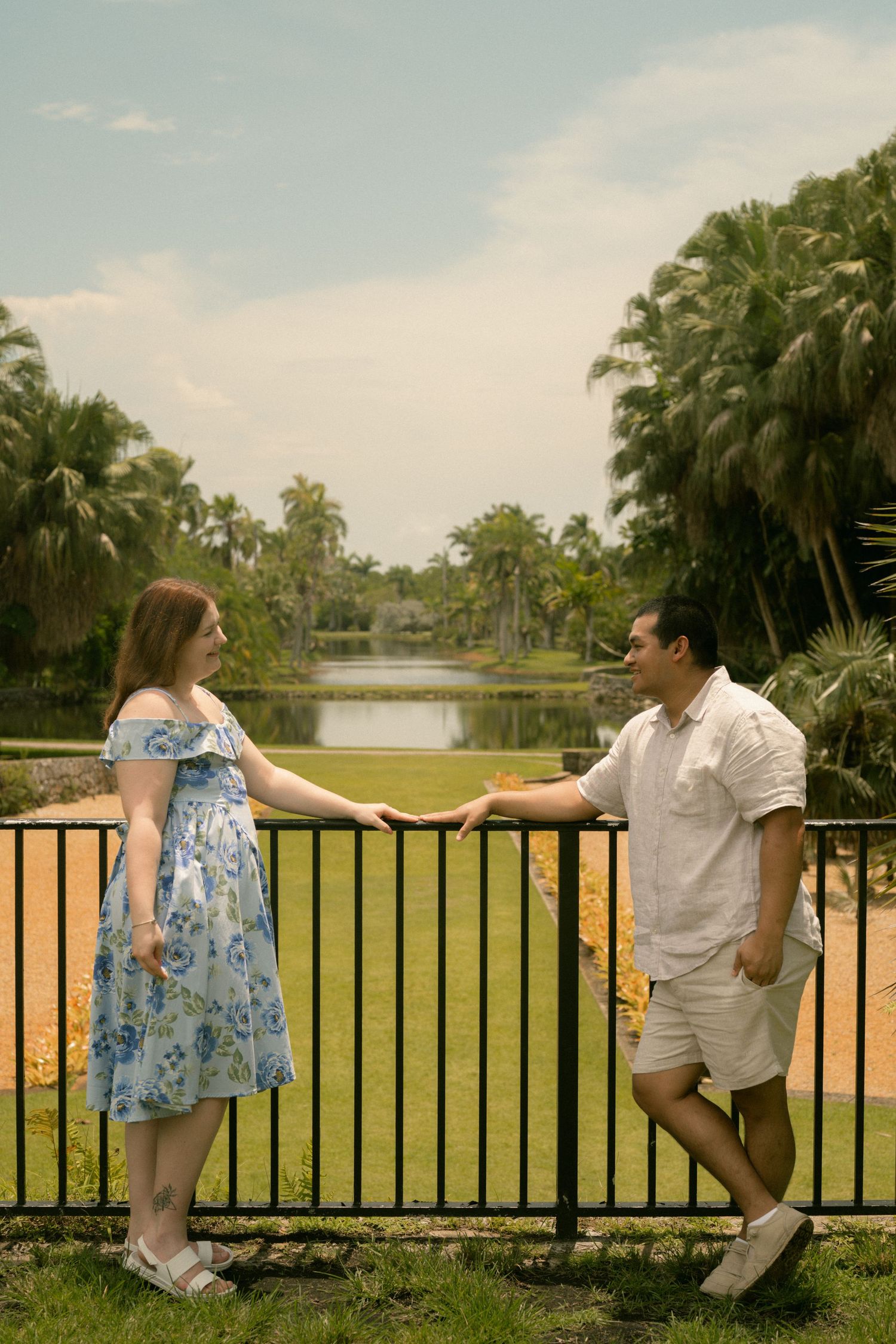 Couple holding hands by a decorative fence in a lush tropical park with a serene lake and palm trees.