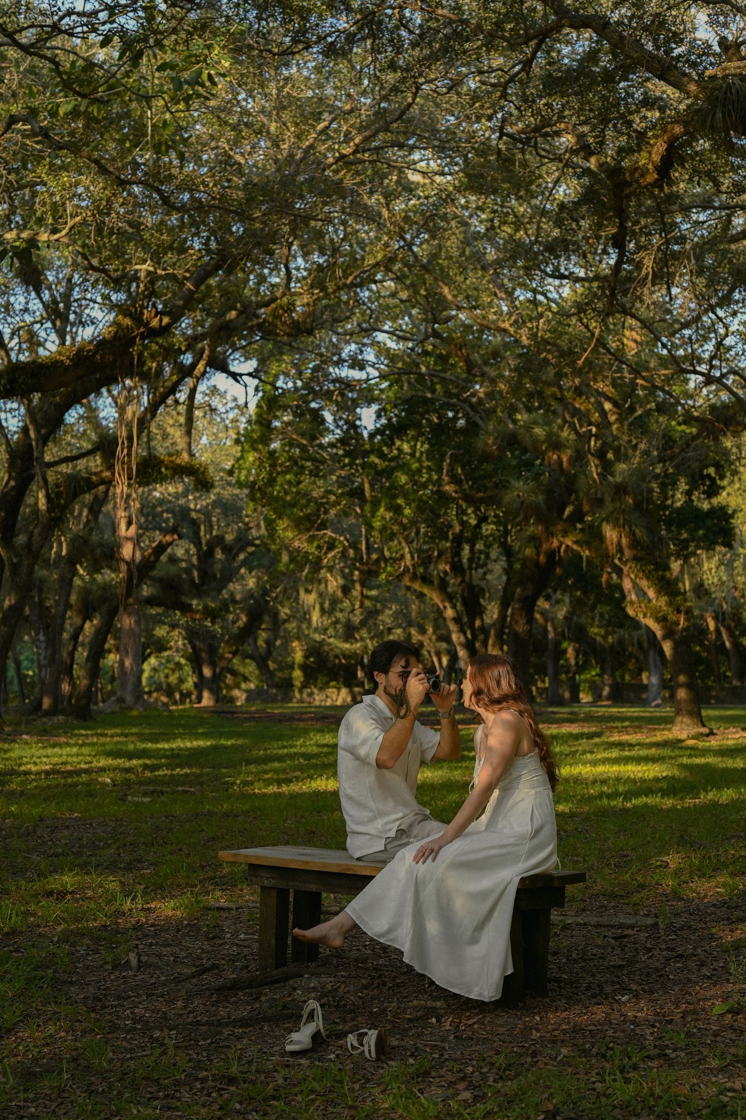 A couple in white attire sits together on a wooden bench under majestic oak trees in a serene outdoor setting.