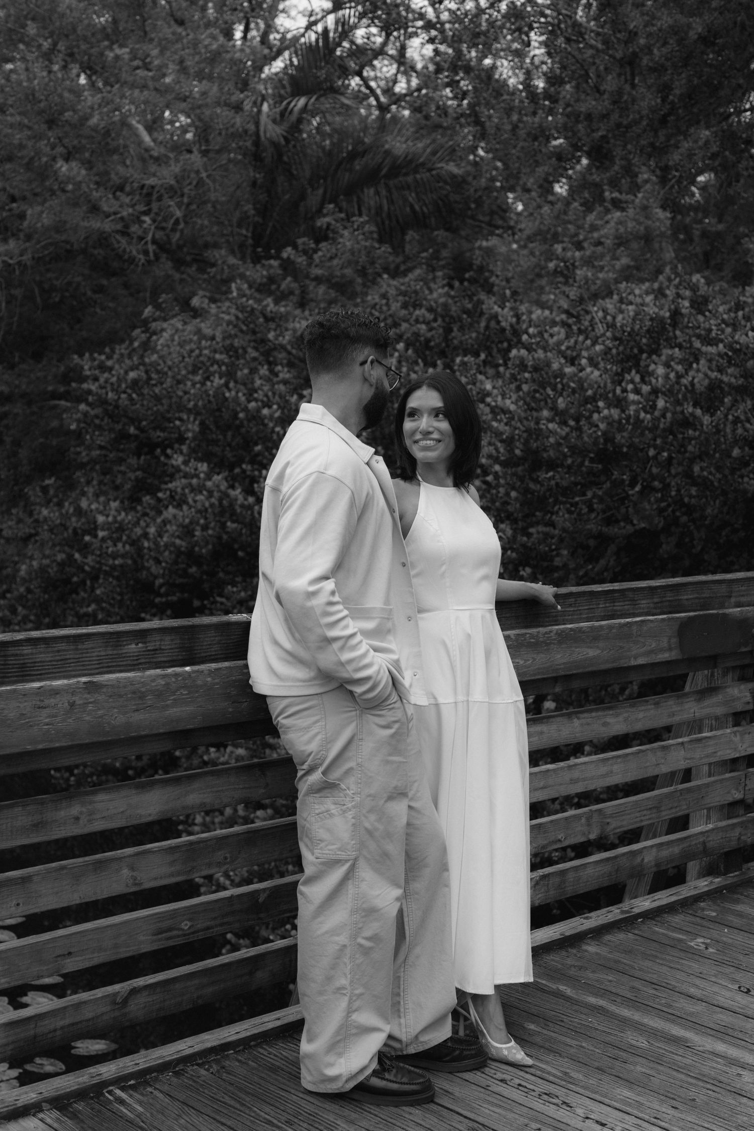 Black and white photo of a couple standing close on a wooden bridge, woman in white dress smiling softly.