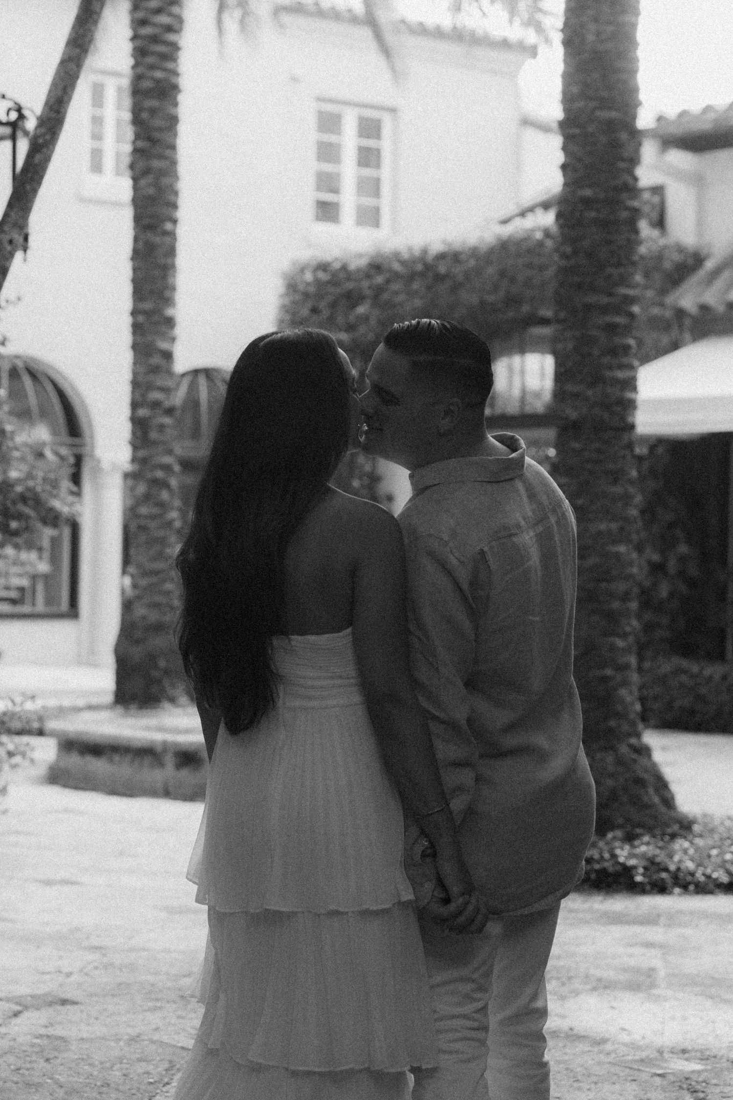 Black and white photo of a couple kissing and holding hands outdoors near palm trees and a building.