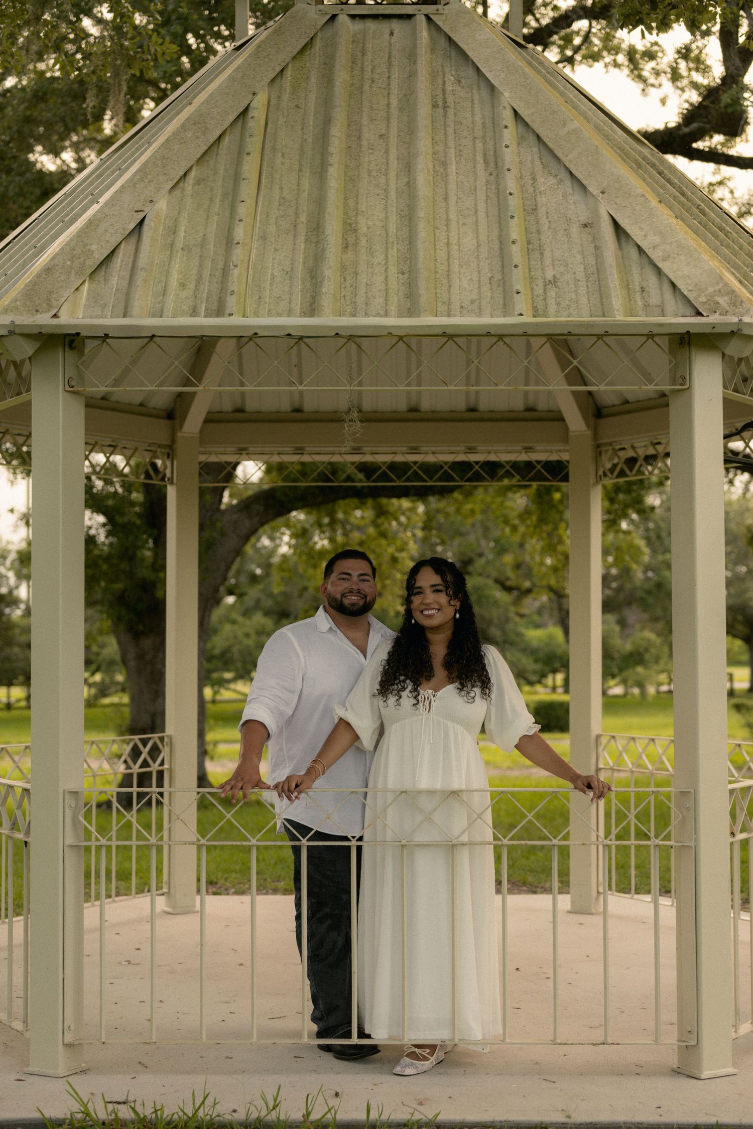 Couple in white outfits posing together inside a charming gazebo in a lush green park setting.
