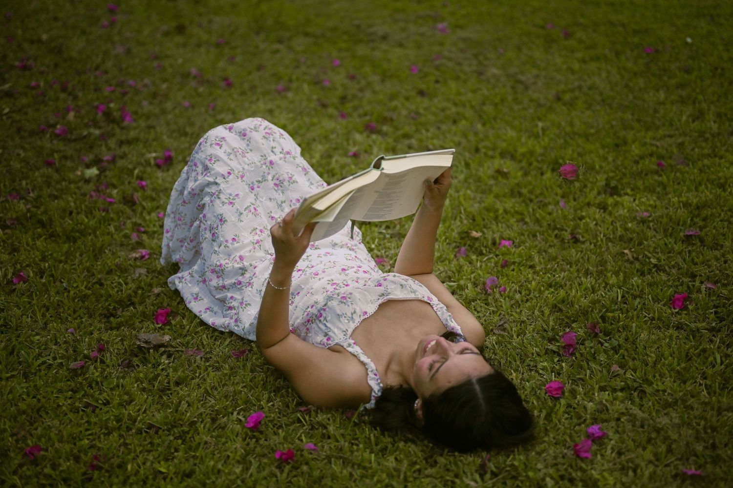 Someone in a floral dress reclines on grass while reading outdoors.
