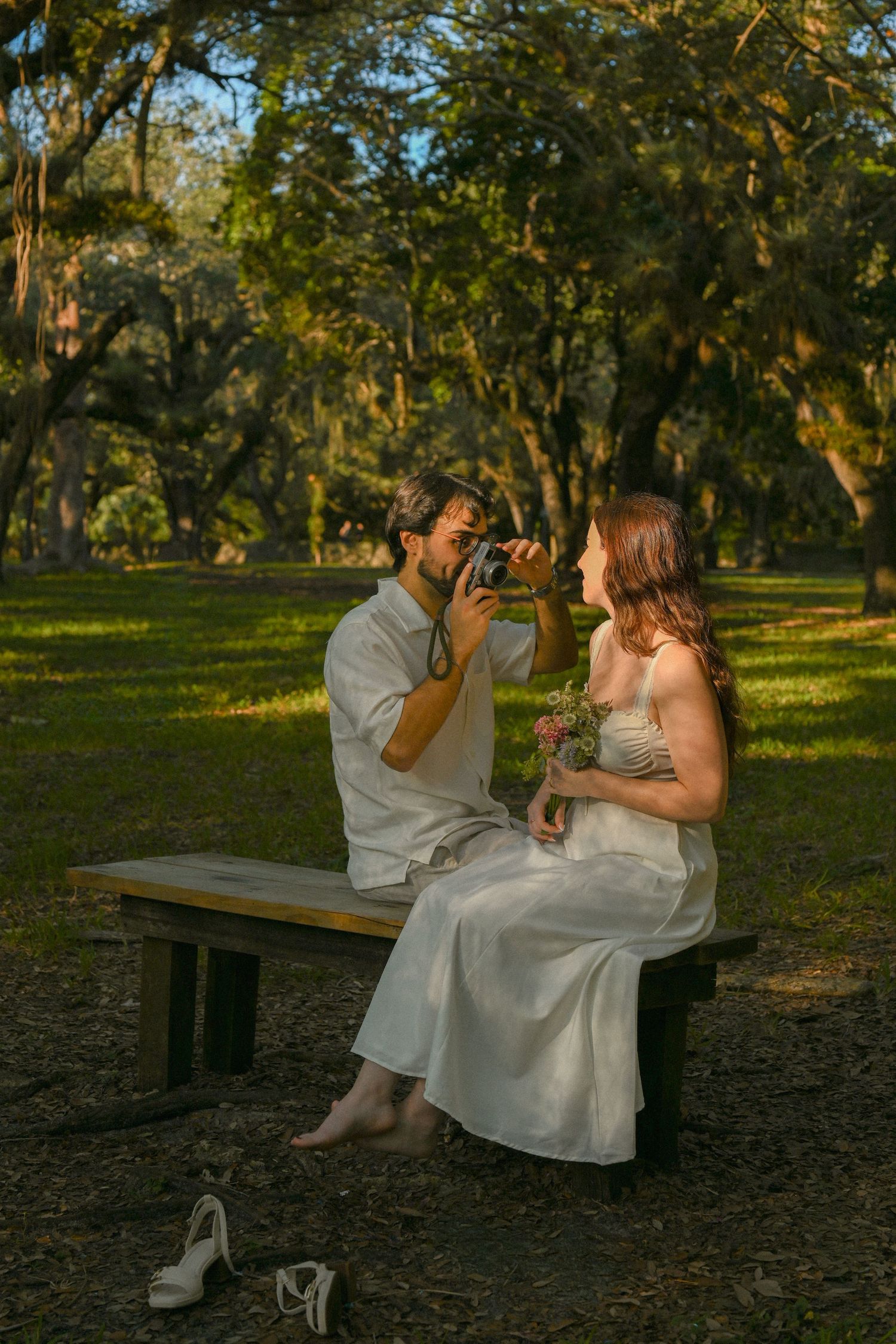 A couple in white outfits shares drinks while sitting on a wooden bench in a sunlit park setting.