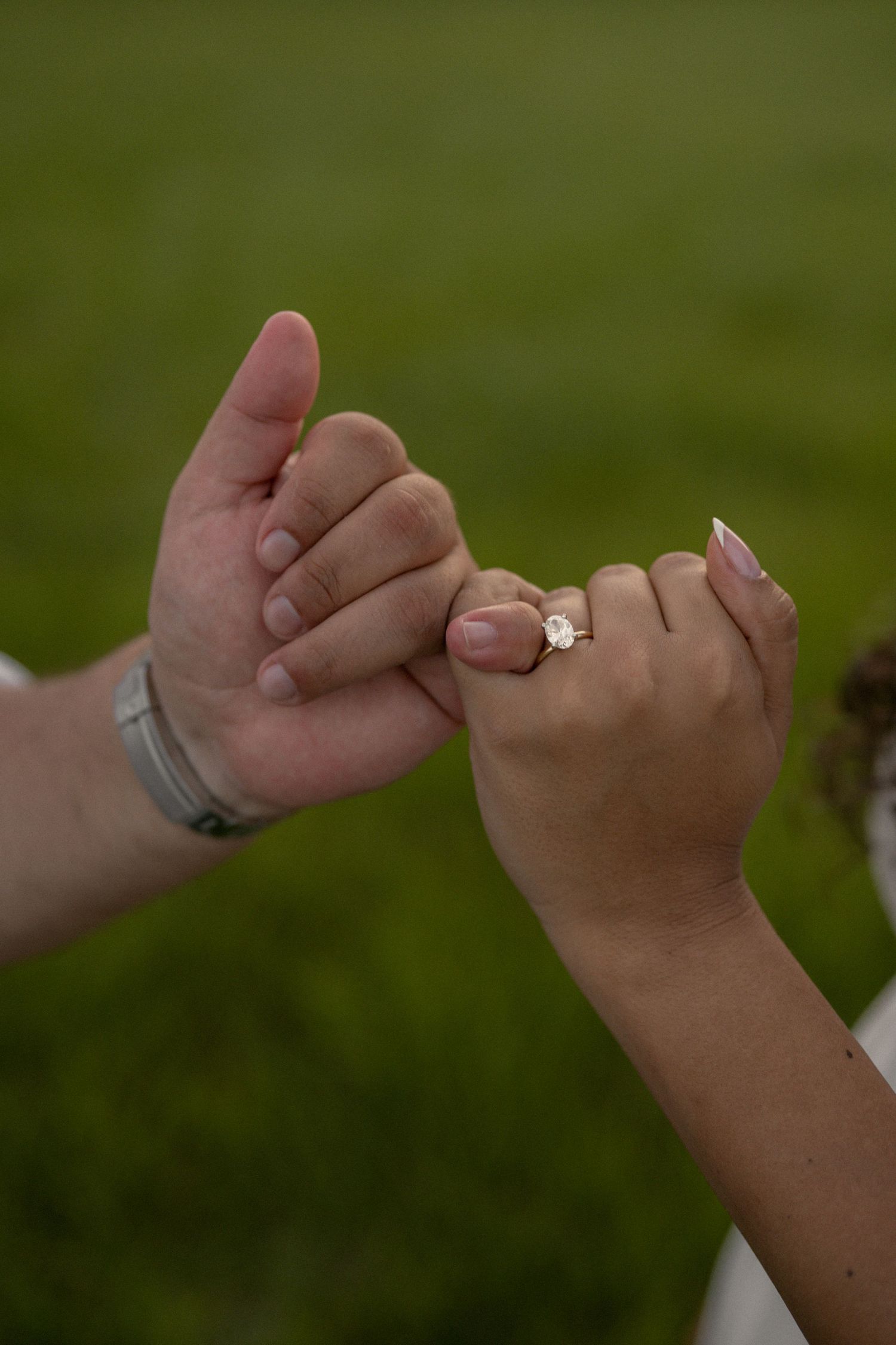 Two hands doing a pinky promise, with the woman wearing a diamond engagement ring, set against a green background.