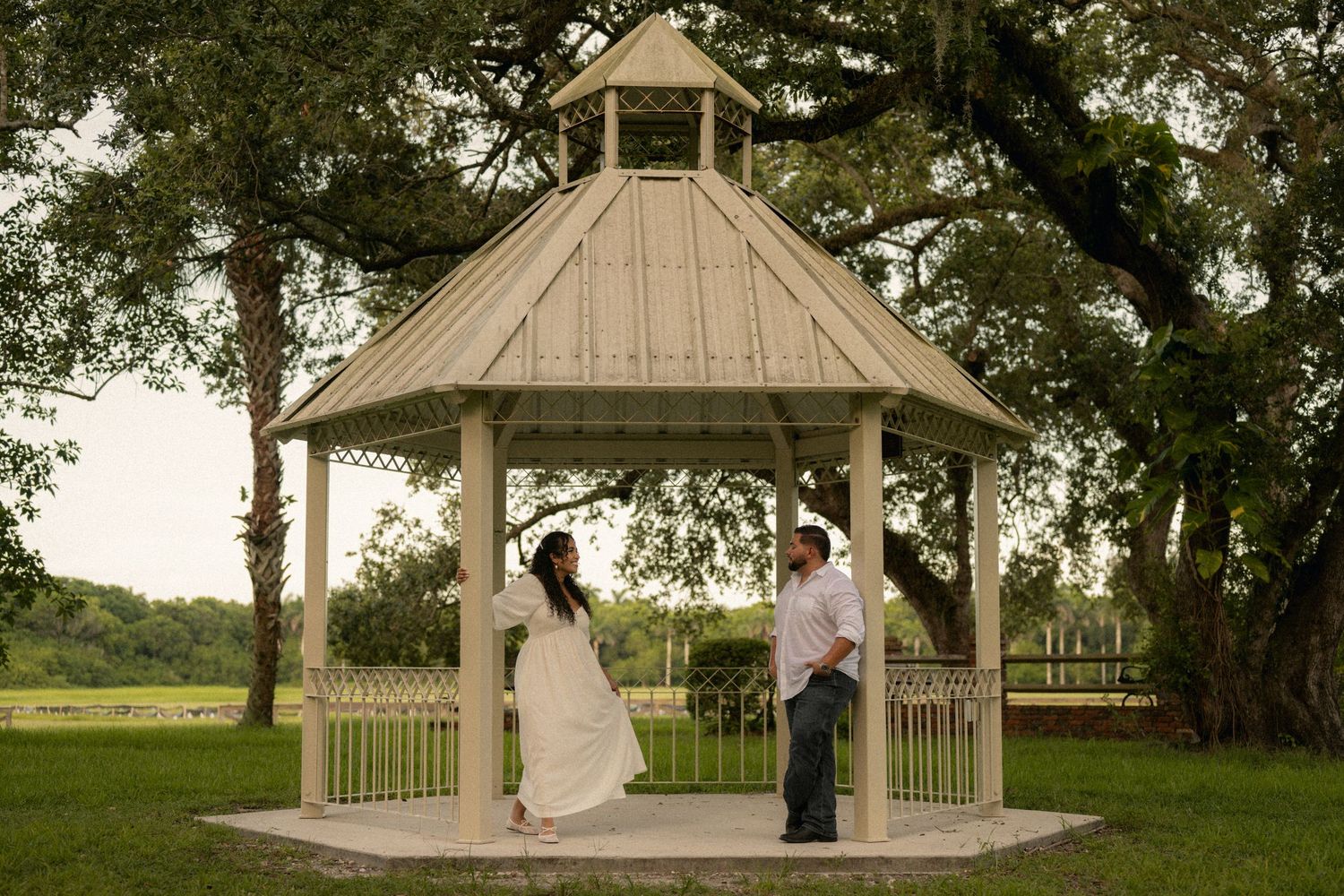 Couple standing together under a white wooden gazebo surrounded by lush green trees on a sunny day.