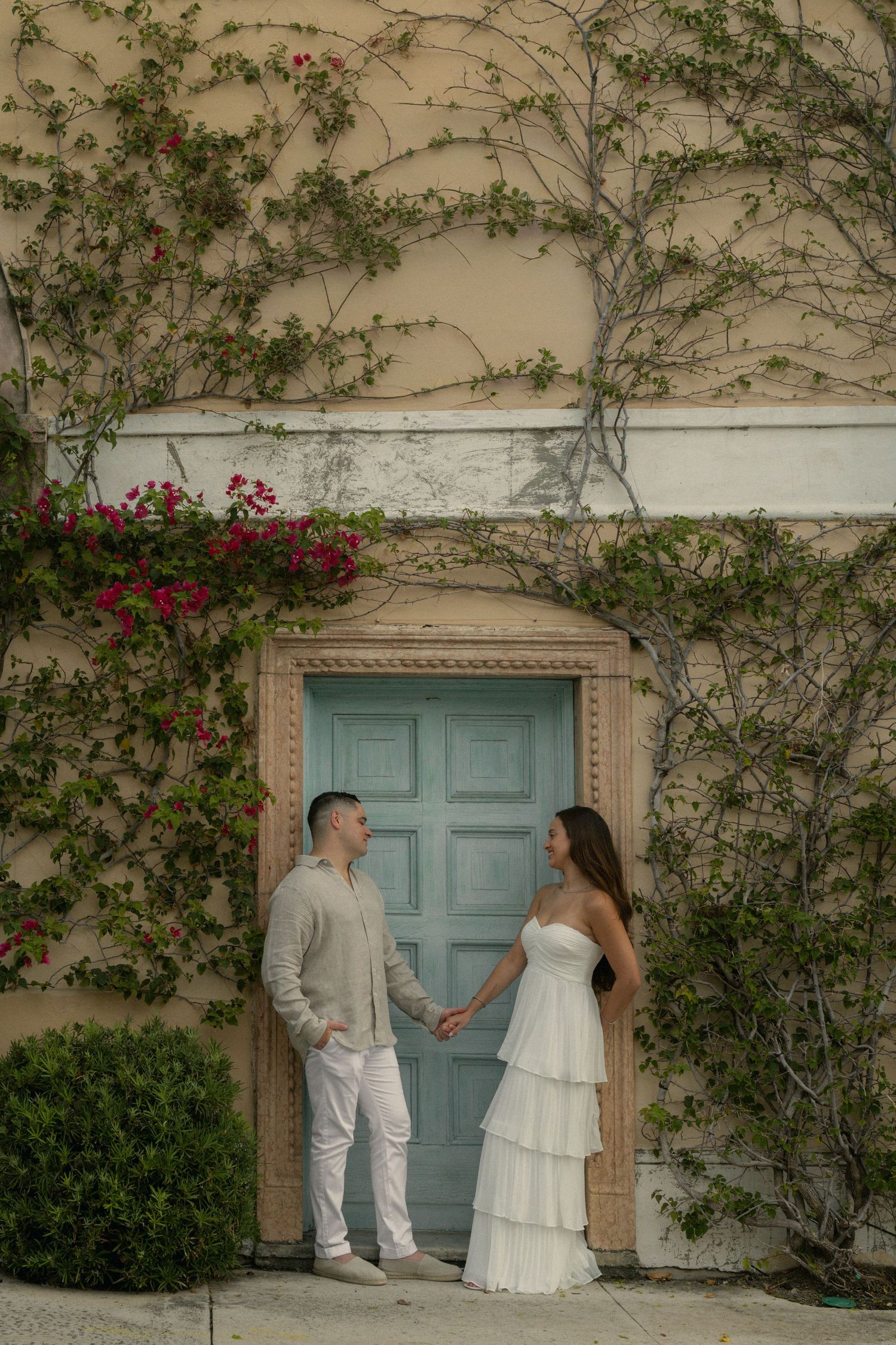 Couple holding hands in white outfits before a teal door, framed by ivy and bougainvillea on a beige wall.