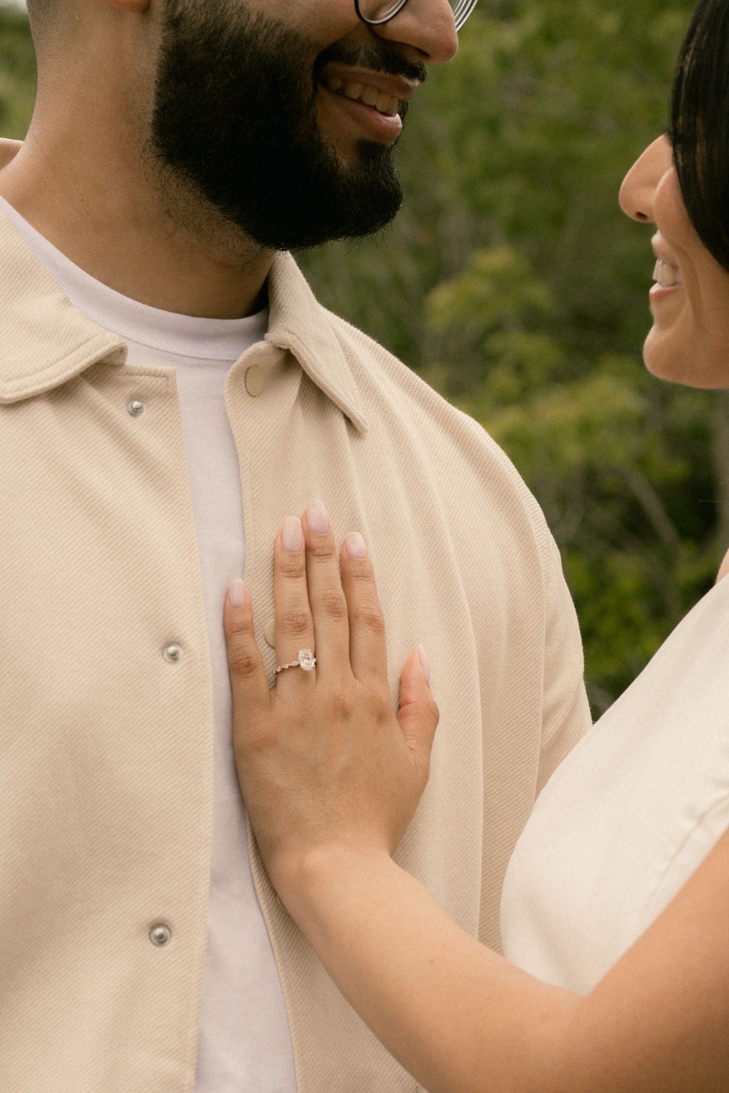 Smiling couple outdoors, woman wearing diamond engagement ring rests hand on man's chest in beige shirt.