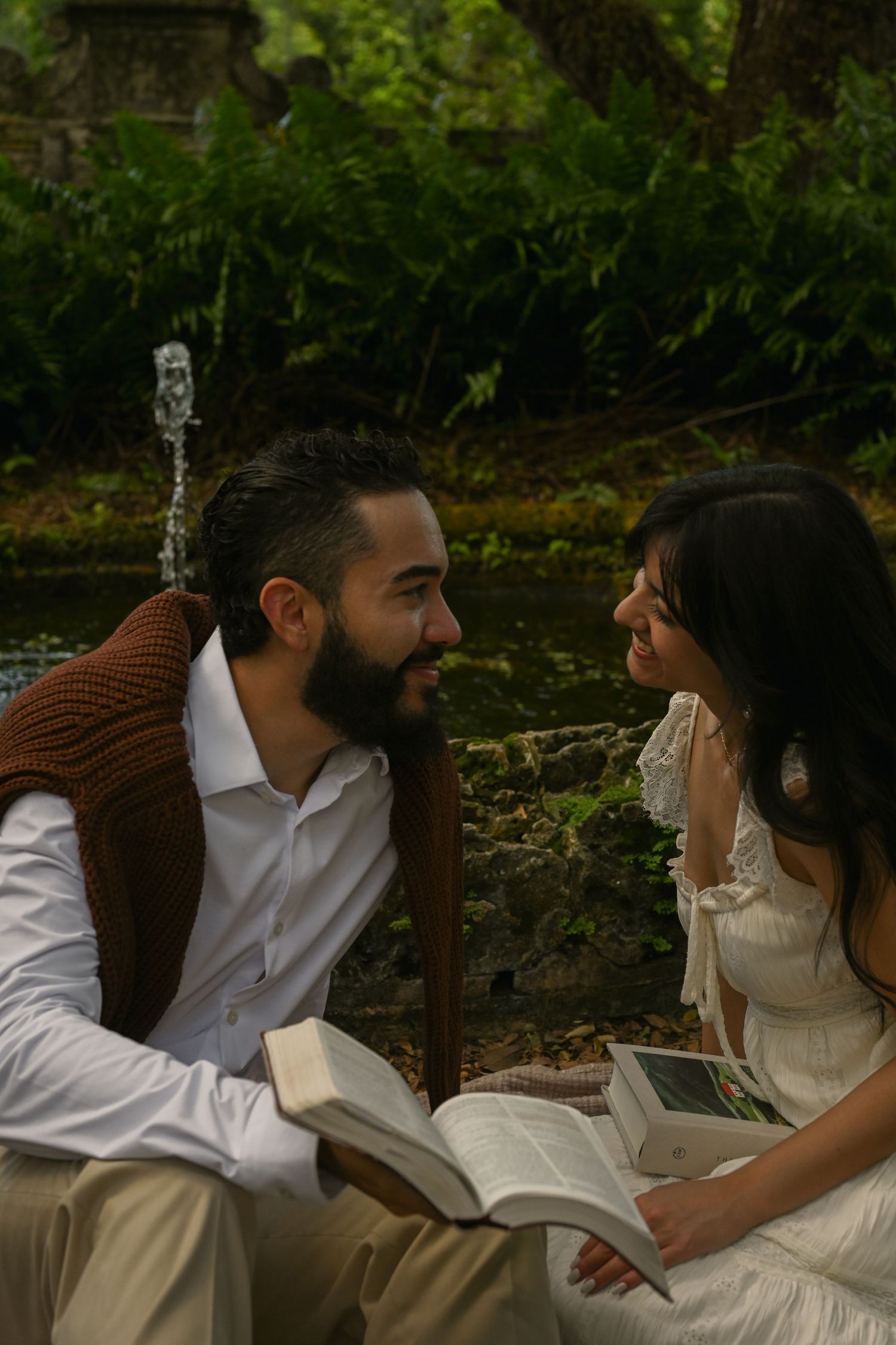 A couple in white sits together on a rustic wooden bench in a garden setting.