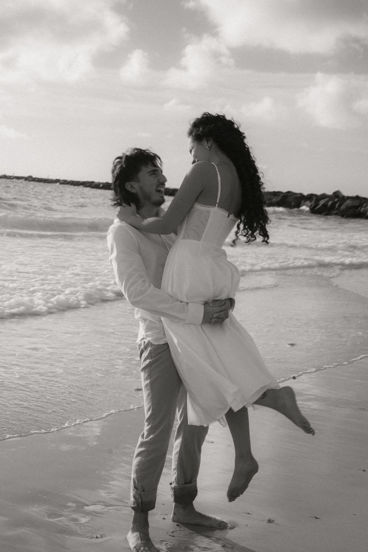 Black and white photo of a couple embracing on a beach, man lifting woman in white dress by ocean waves.
