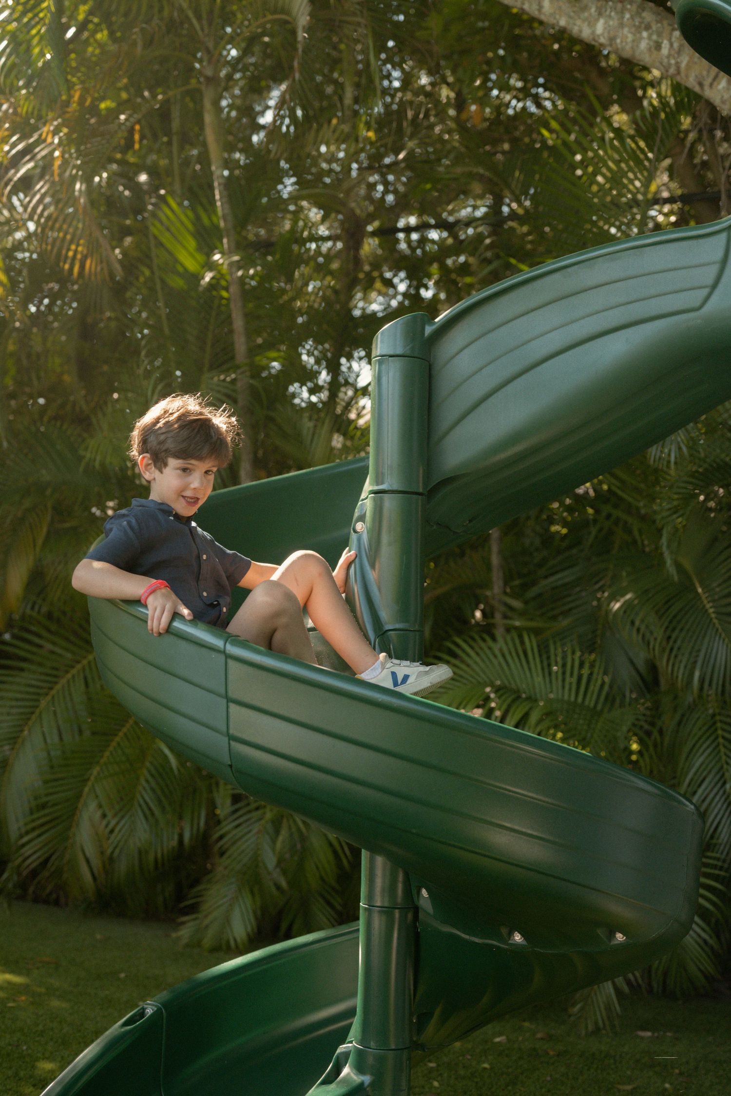 A smiling boy in a dark shirt sits on a green spiral slide surrounded by lush tropical palms on a sunny day at the playground.
