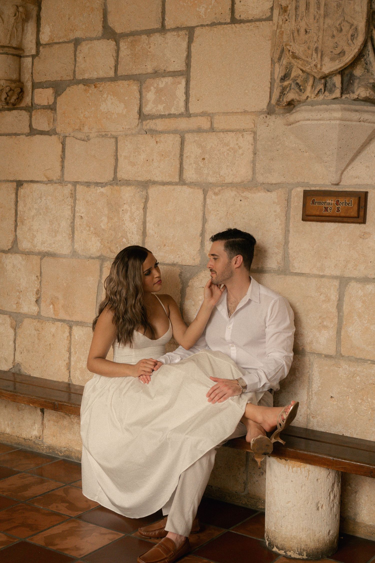 Couple in white outfits sit close on a wooden bench against a limestone wall, sharing a tender moment in a historic stone cloister.
