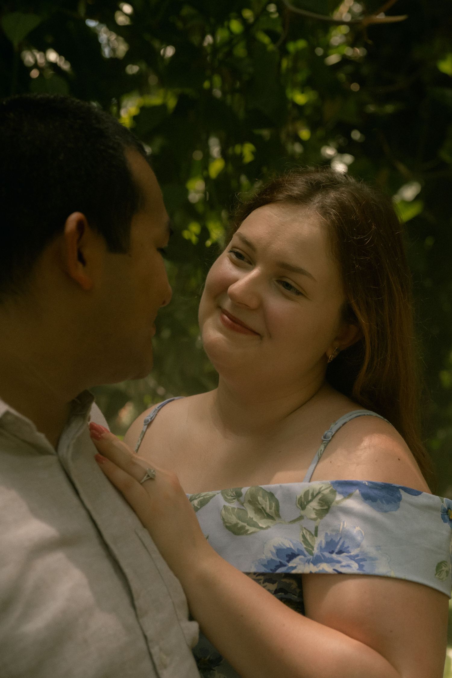 A young couple shares a tender moment outdoors, with the woman smiling warmly in a floral off-shoulder dress amid lush greenery.