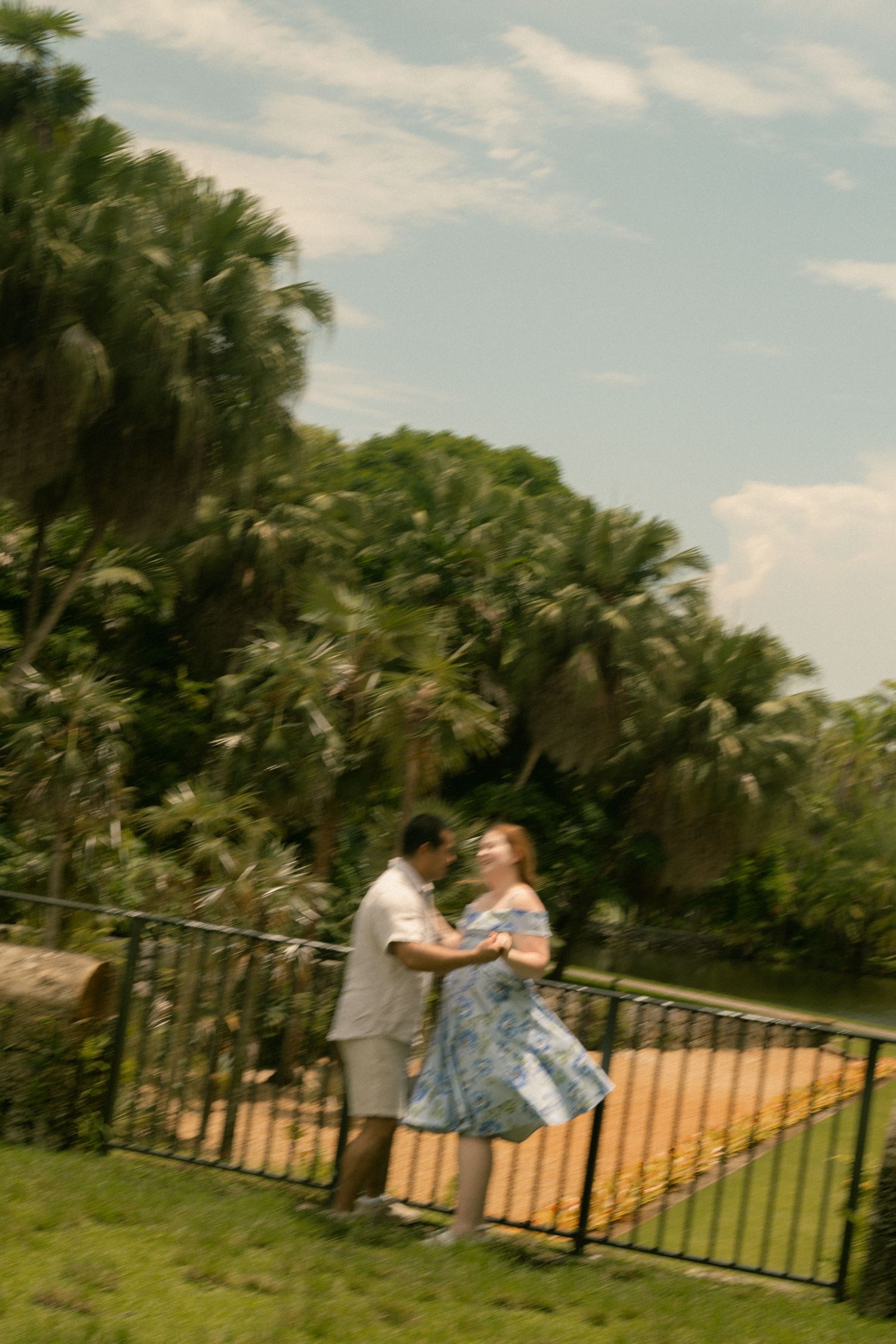 Couple dancing together by a black iron fence surrounded by lush tropical palm trees under a partly cloudy sky.